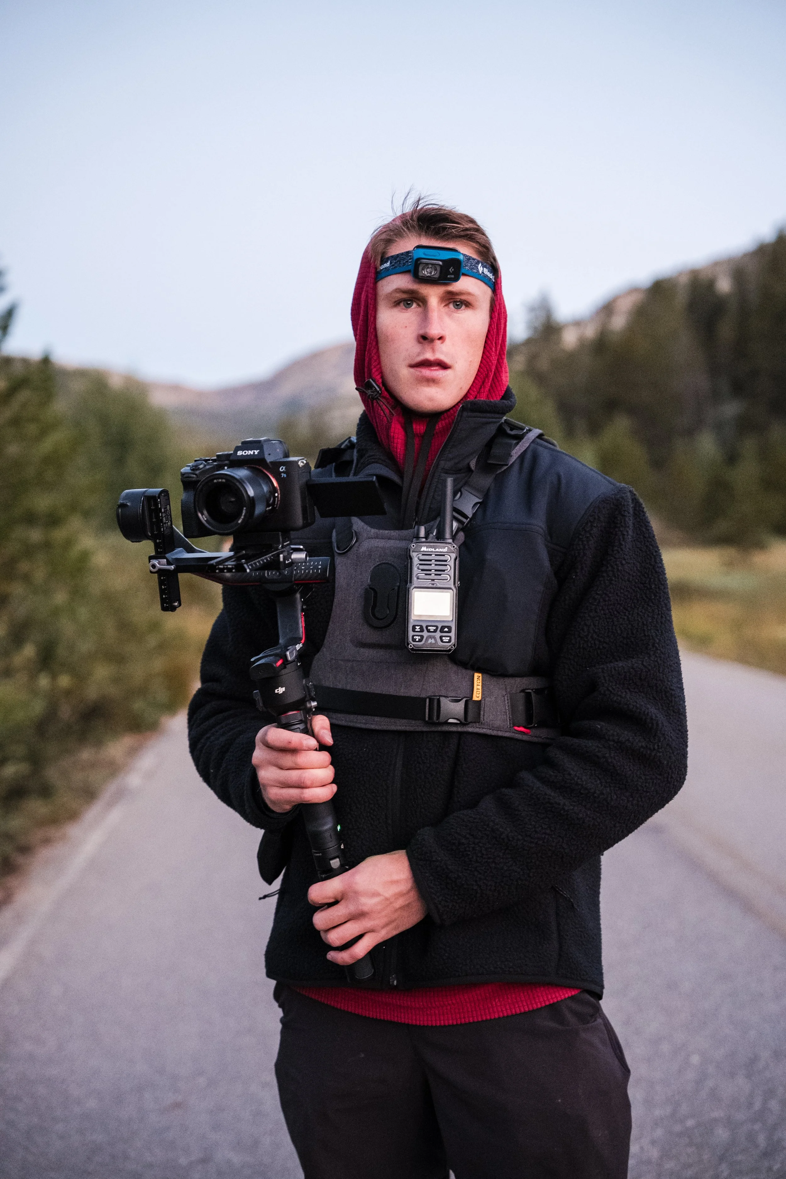 Young man standing outdoors on a path, holding a professional video camera with a stabilizer, wearing a headlamp and outdoor gear, with mountains and trees in the background.