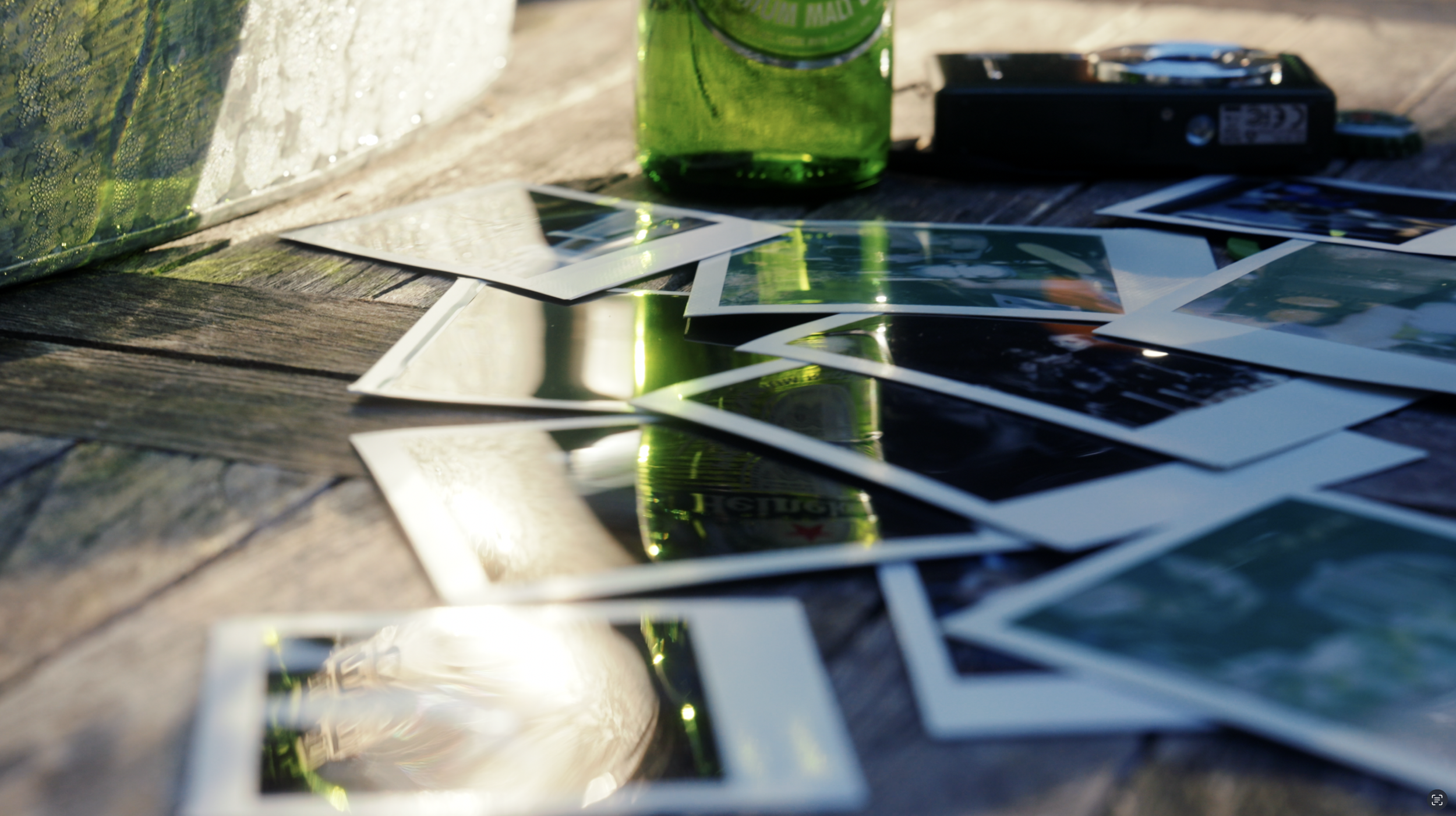 Scattered instant photographs on a wooden surface with a green glass jar and a camera in the background.