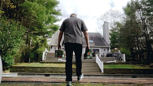 A man walking away from the camera carrying grocery bags toward a house with stairs.