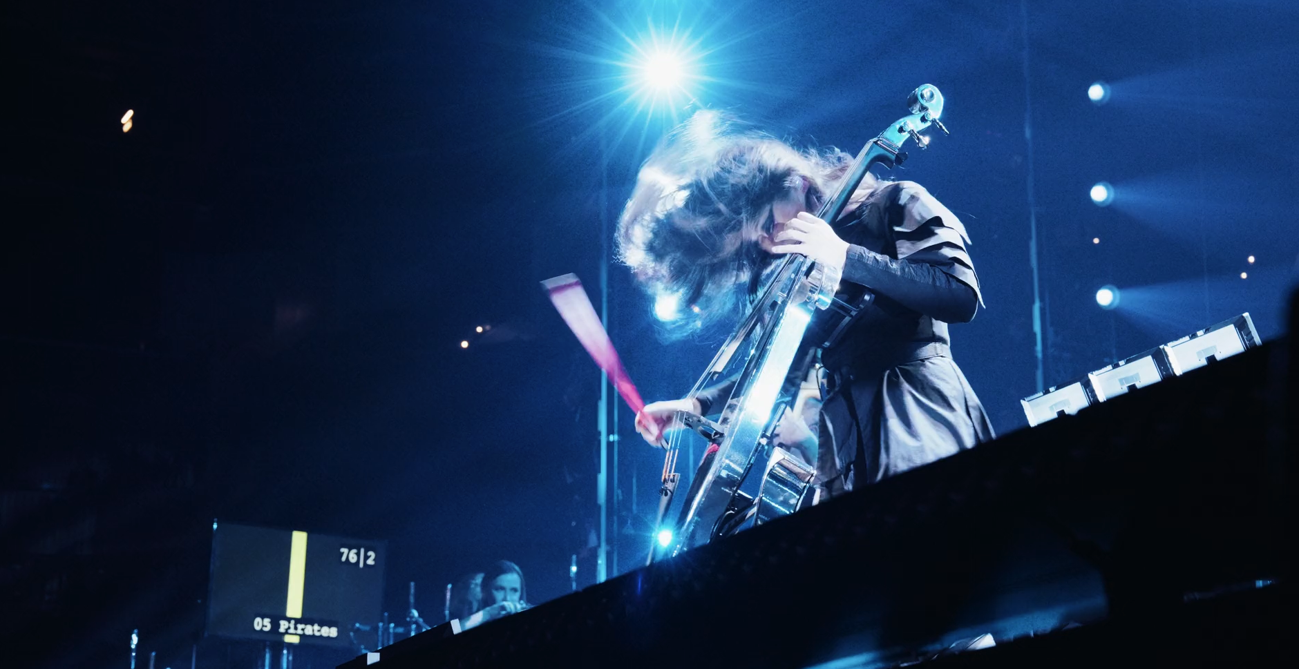 Performer with long hair playing an electric cello on stage, surrounded by blue stage lights and visuals.