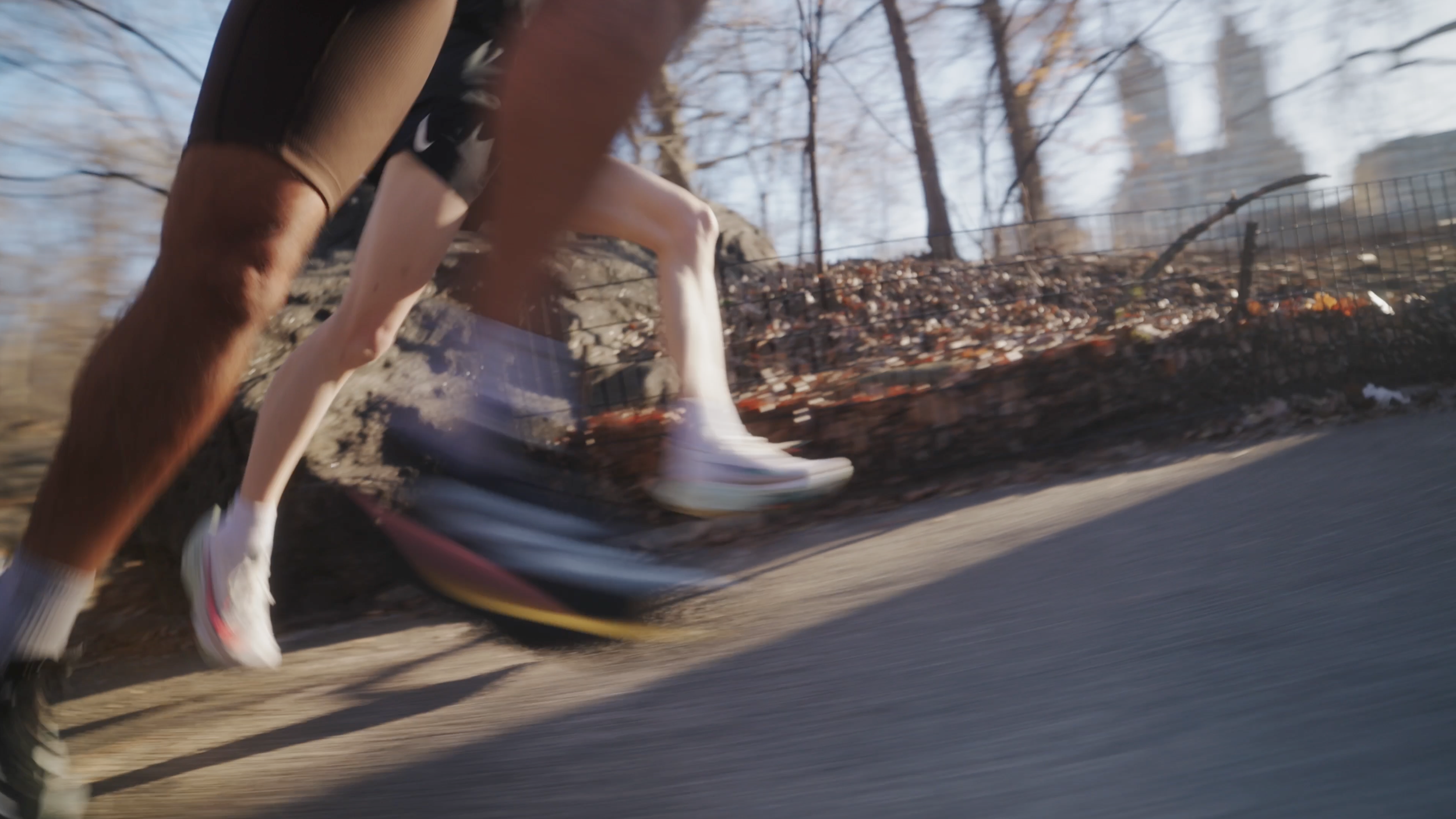 Close-up of two runners' legs and feet running on a paved trail during daytime, with a blurred background of trees and a fence.