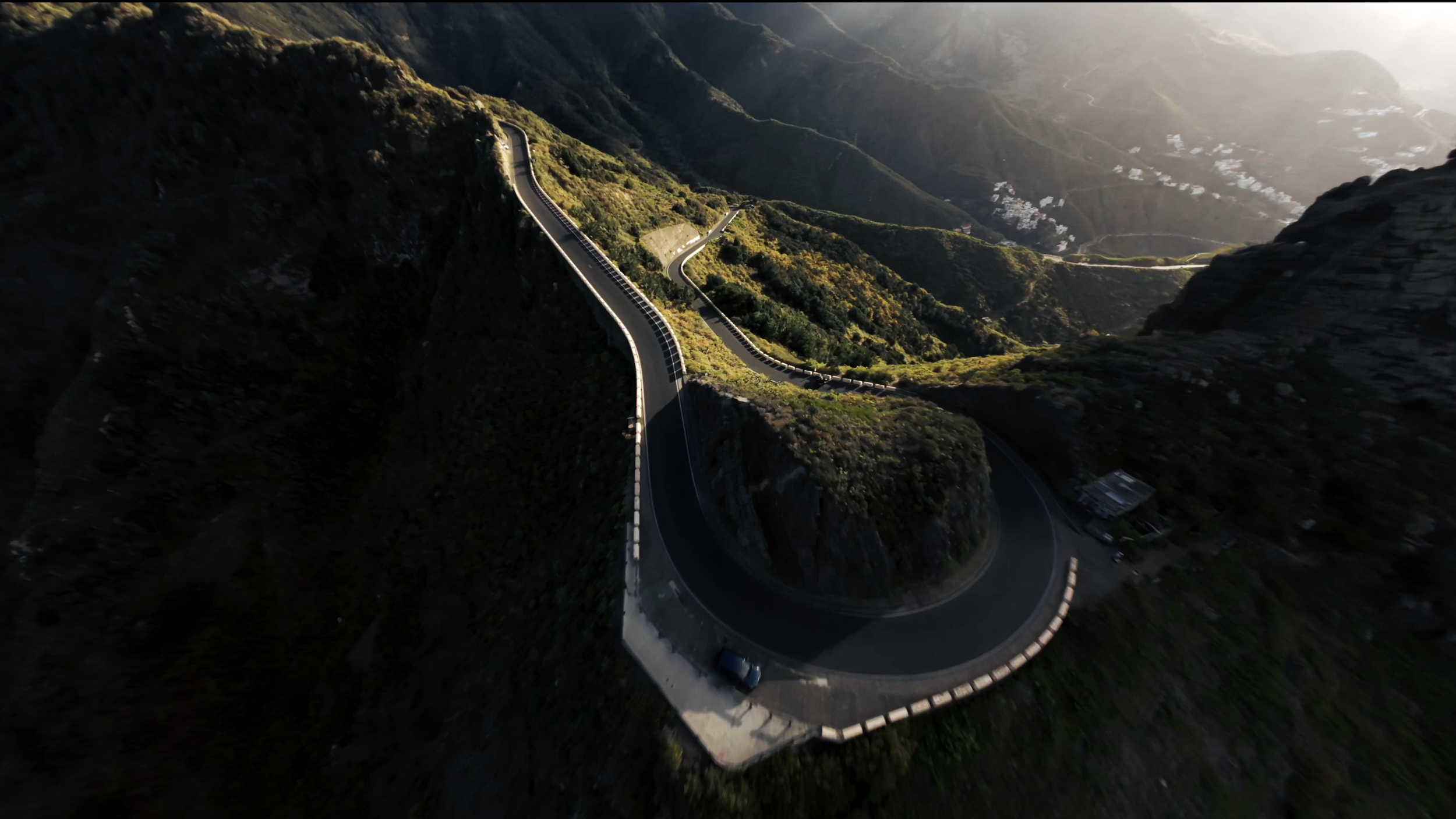 A winding mountain road with sharp curves and guardrails, surrounded by steep hills covered in greenery, with sunlight illuminating parts of the landscape.
