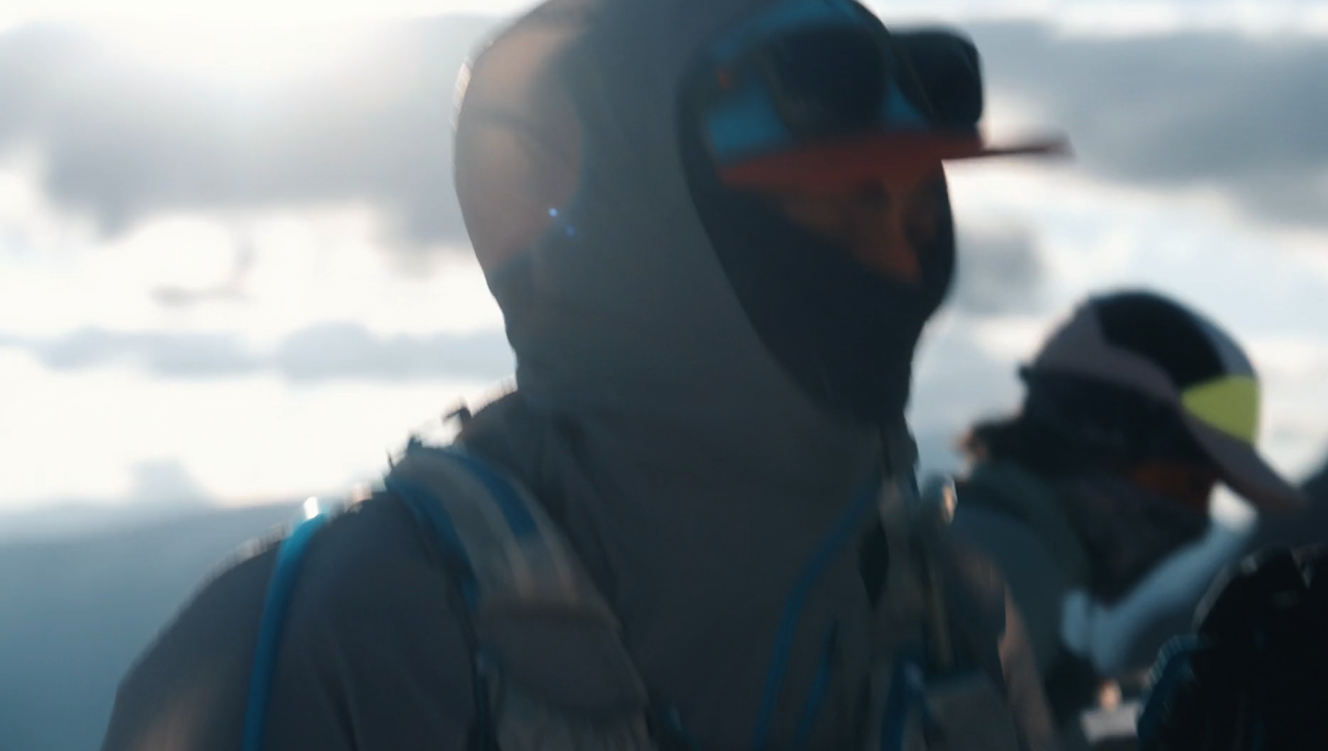 Two people dressed in winter gear, wearing sunglasses and helmets, standing outdoors in a cloudy environment.