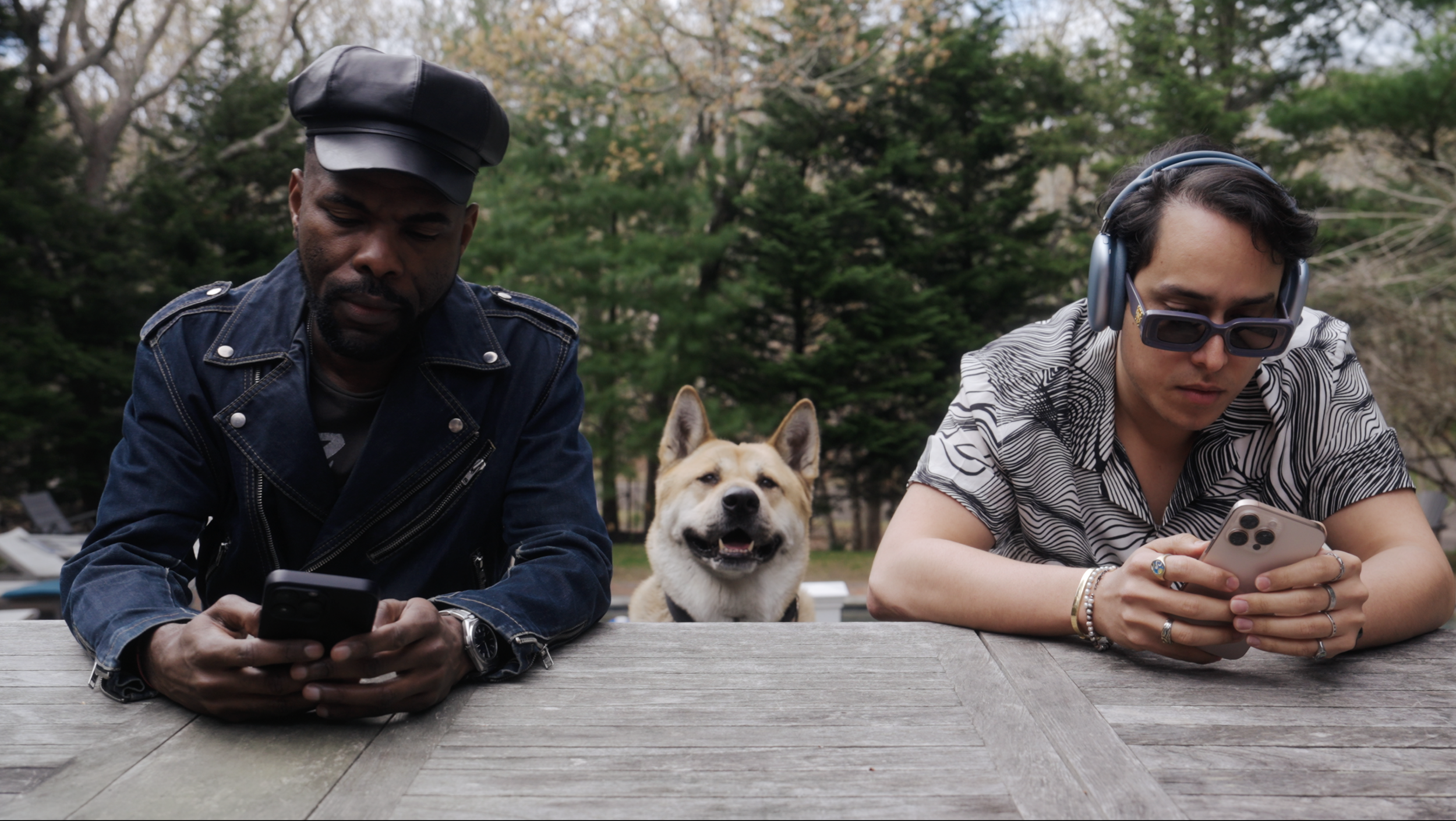 Two men sitting at a picnic table outdoors, one wearing a leather cap and phone, the other with headphones and sunglasses looking at a phone, with a happy dog between them.