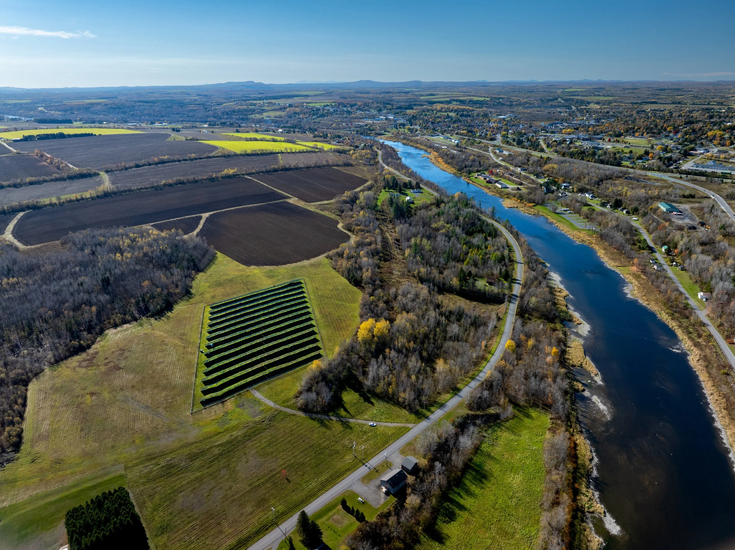 Aerial view of farmland, a river, and a small residential area with roads, trees, and open fields in a rural landscape.
