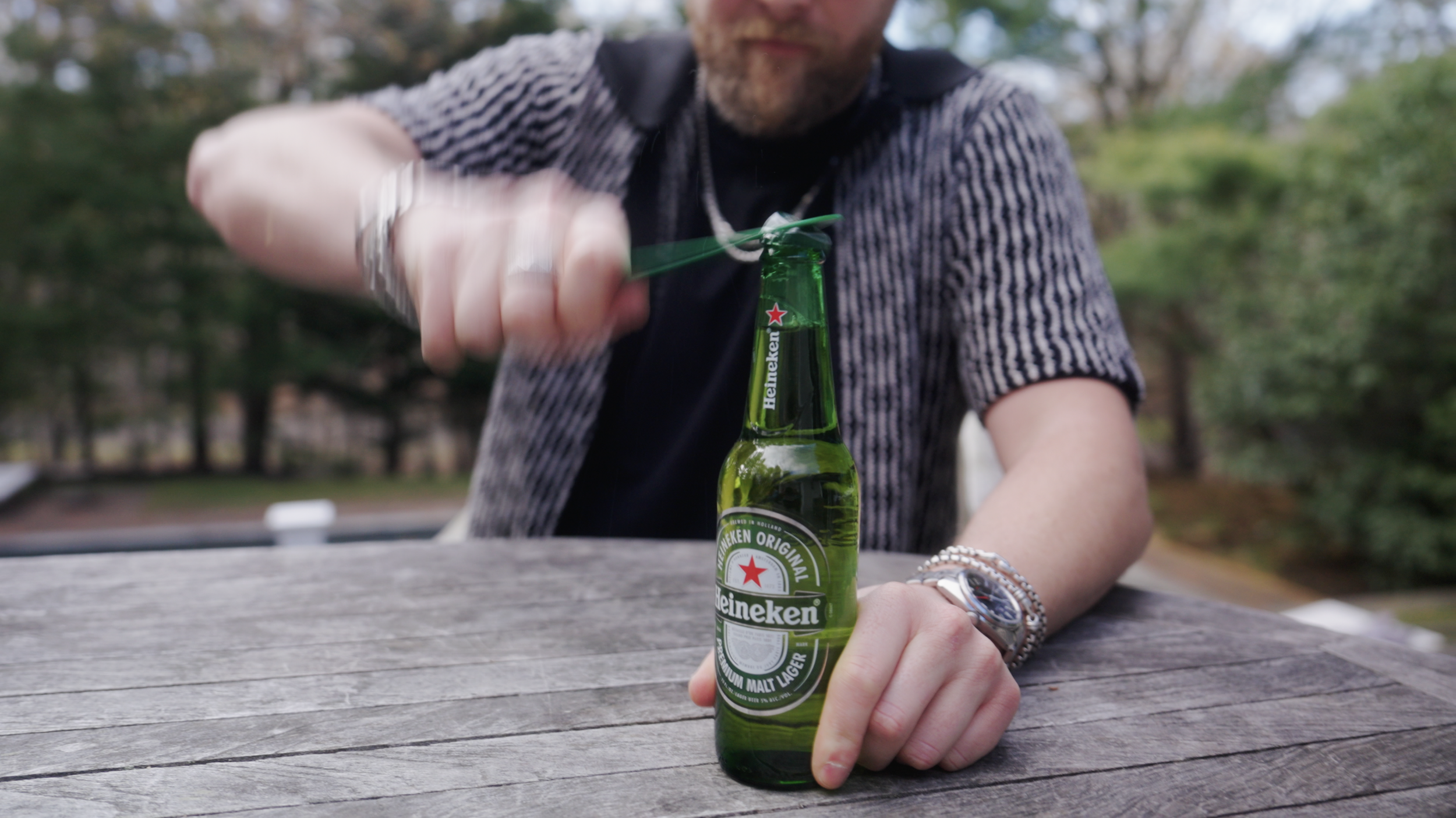 A man is opening a Heineken beer bottle with a bottle opener outdoors, sitting at a weathered wooden table with green trees and a park in the background.