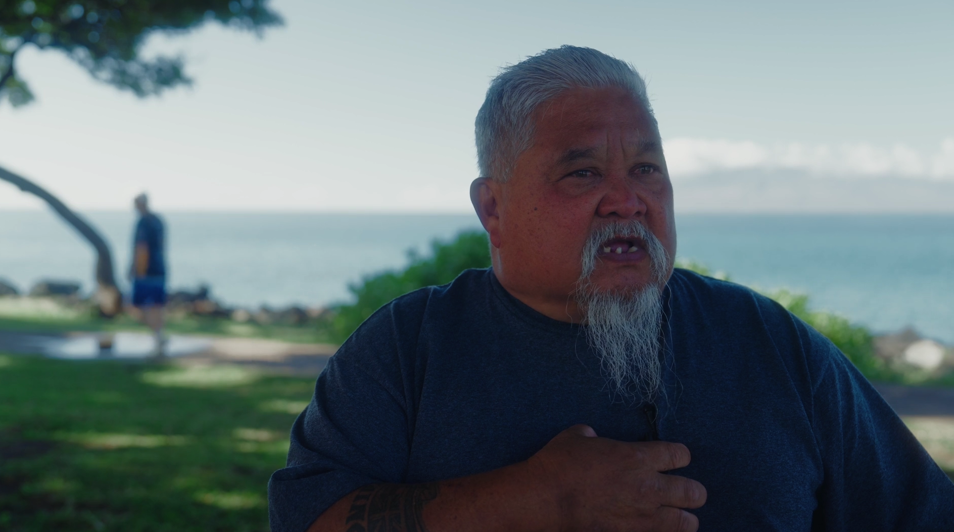 An older man with gray hair, a beard with a mustache, and missing teeth, wearing a navy blue shirt, sitting outdoors near the coast with trees and a person in the background.