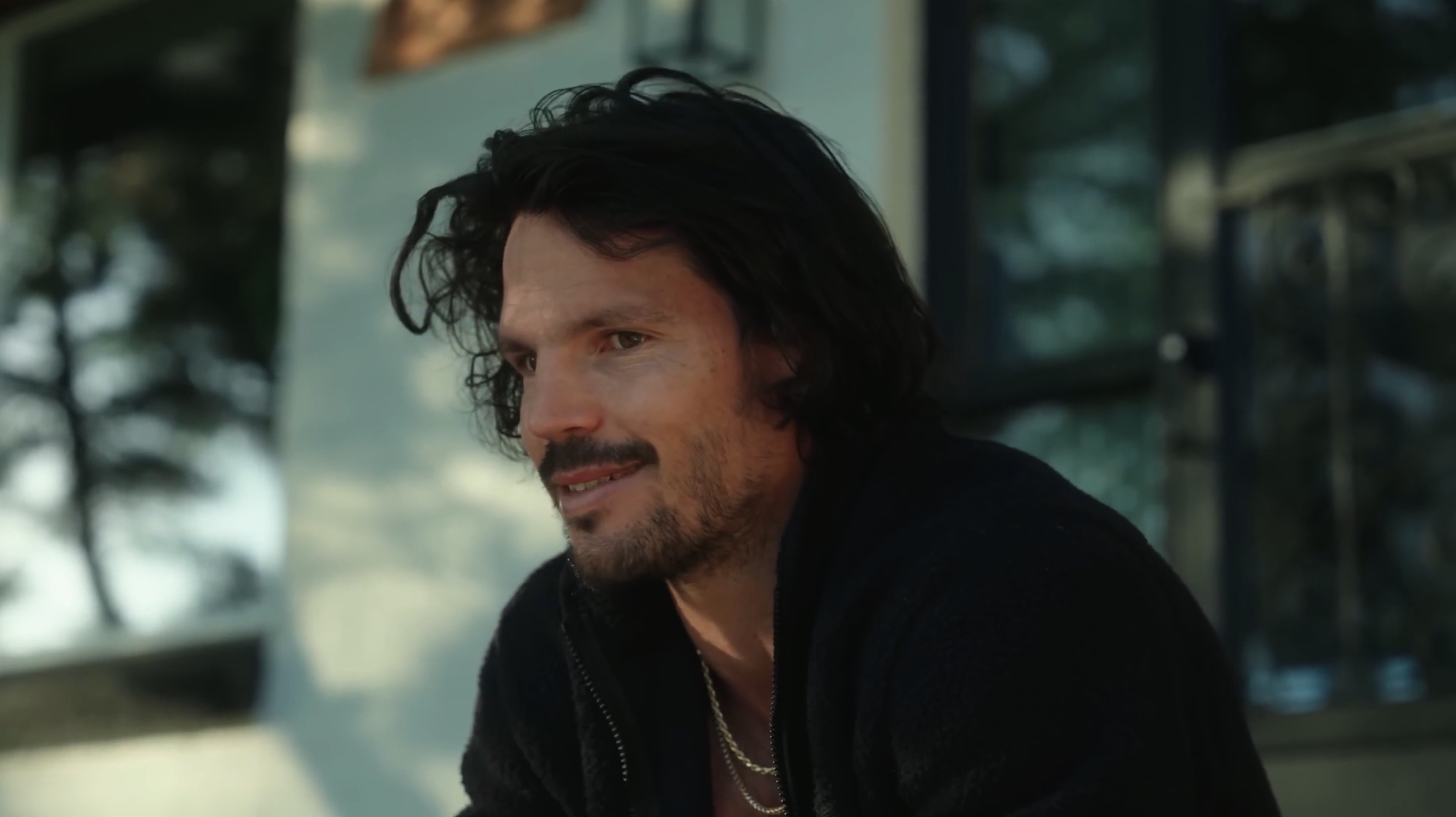 A man with dark, curly hair and a beard is smiling while sitting outdoors, with trees and a building reflection in the background.