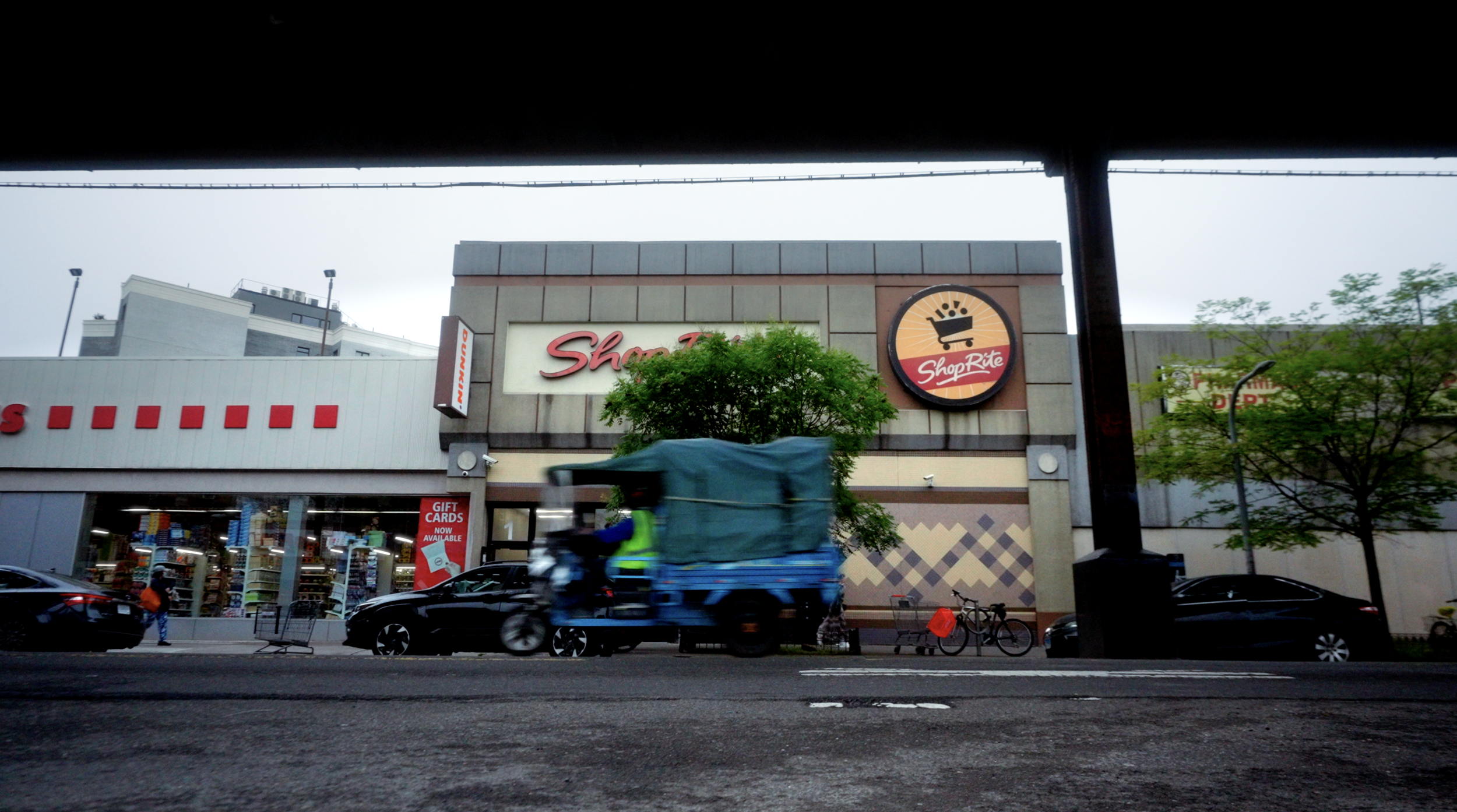 View of a shopping center with a storefront labeled ShopRite, a logo, trees, parked cars, a bicycle, and a delivery vehicle passing by, under an overpass.