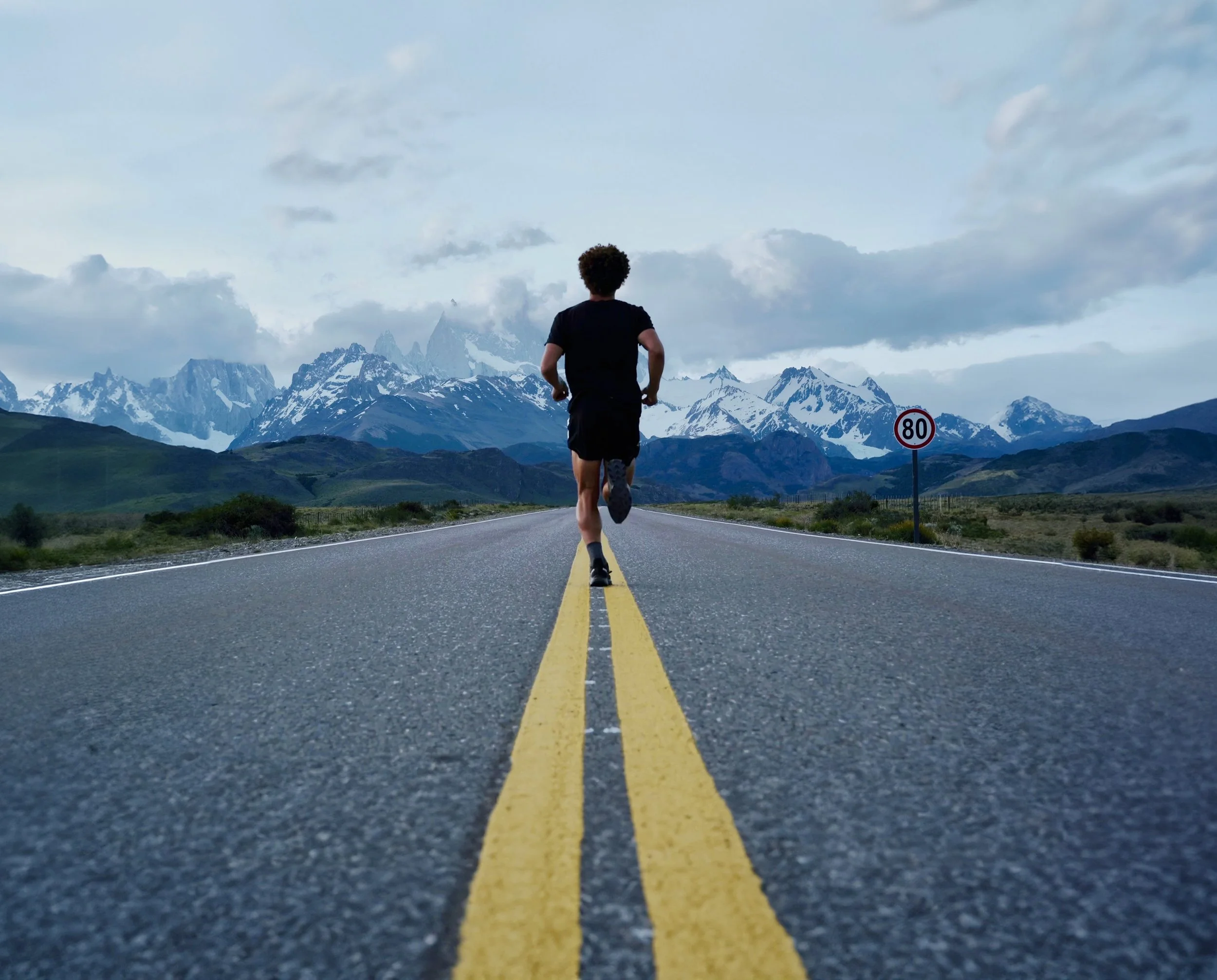 A man running on a road with mountains in the background, a speed limit sign showing 80 km/h, and cloudy sky.