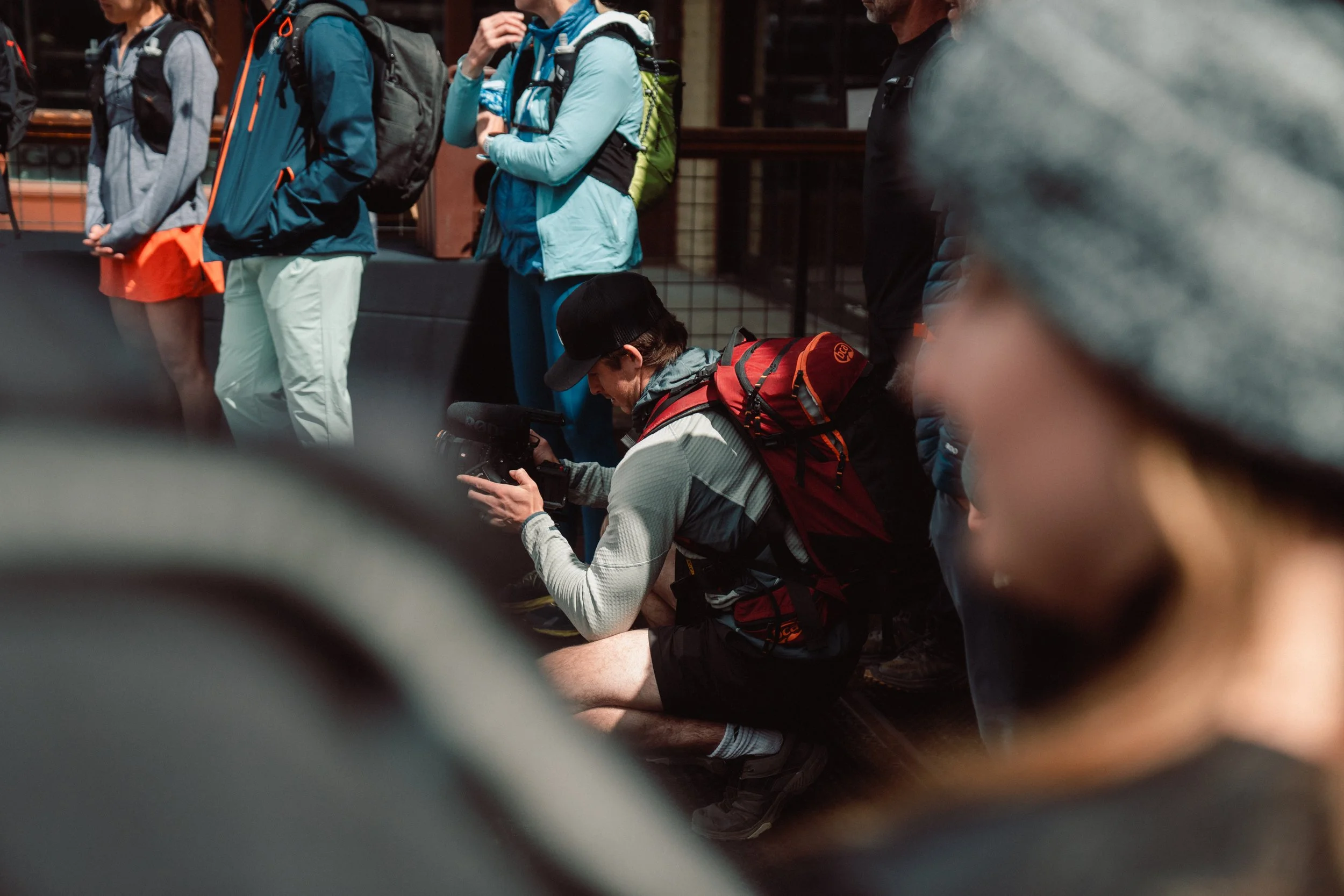 A group of people, mostly in athletic gear and backpacks, are standing and kneeling indoors, with one person sitting on the floor using a camera or recording device.