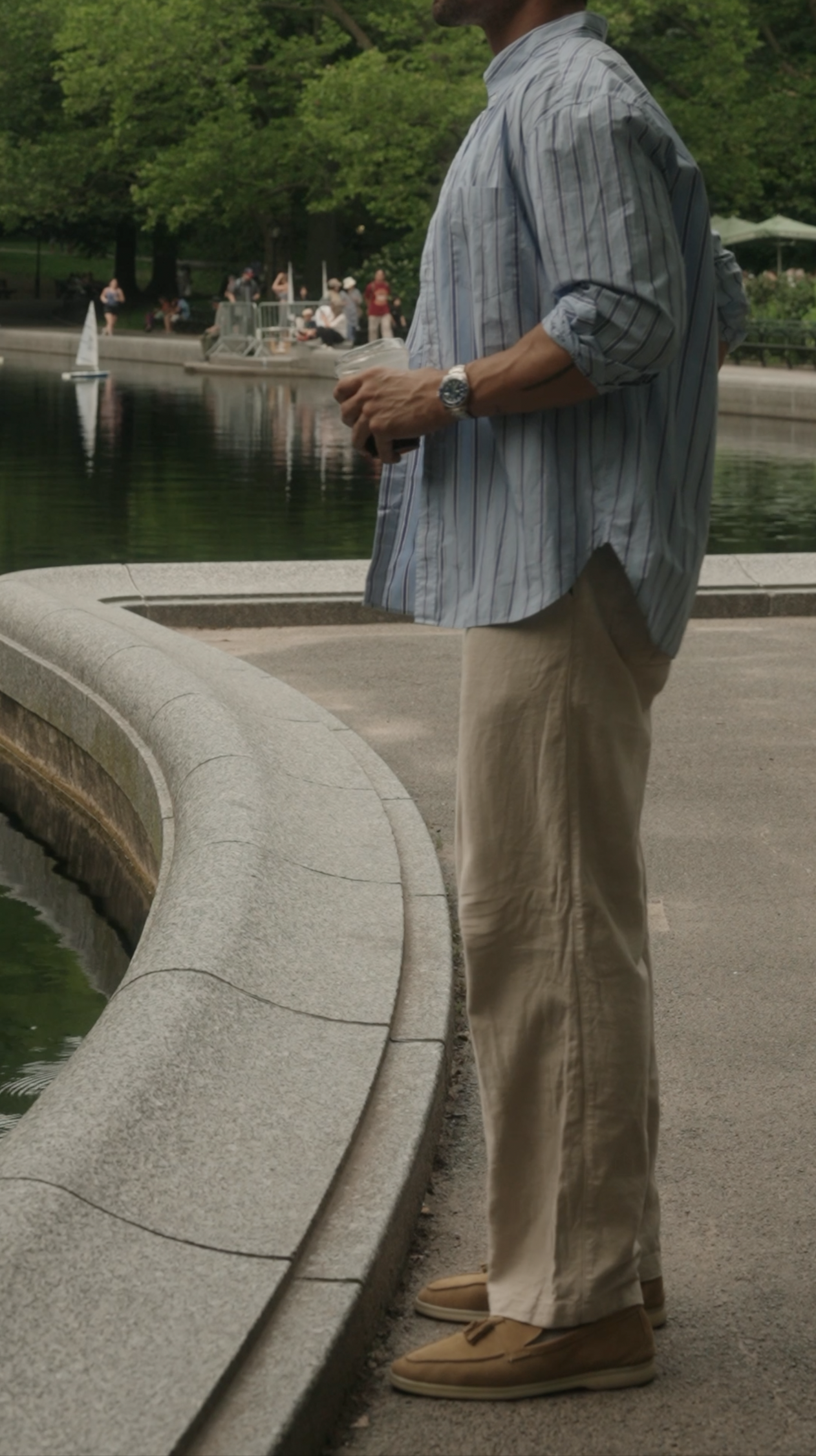 Man standing by a park pond holding a cup, wearing a striped shirt, beige pants, and tan shoes, with trees and people in the background.