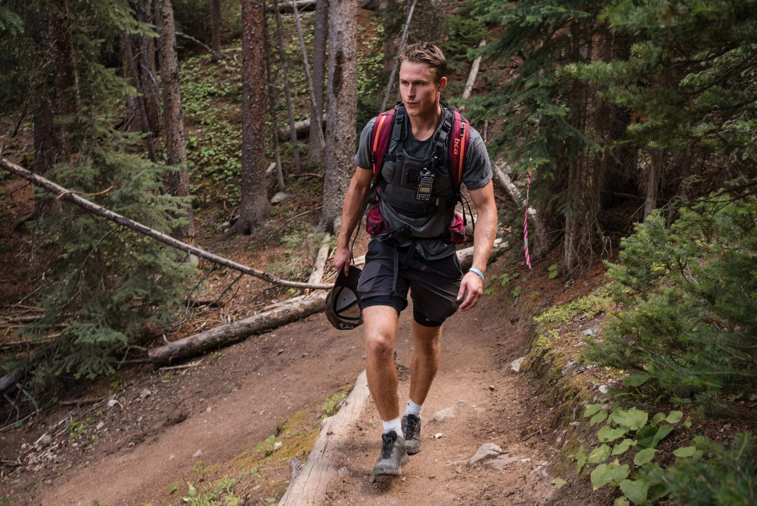 A young man hiking on a dirt trail in a forest, carrying a backpack and holding a helmet.