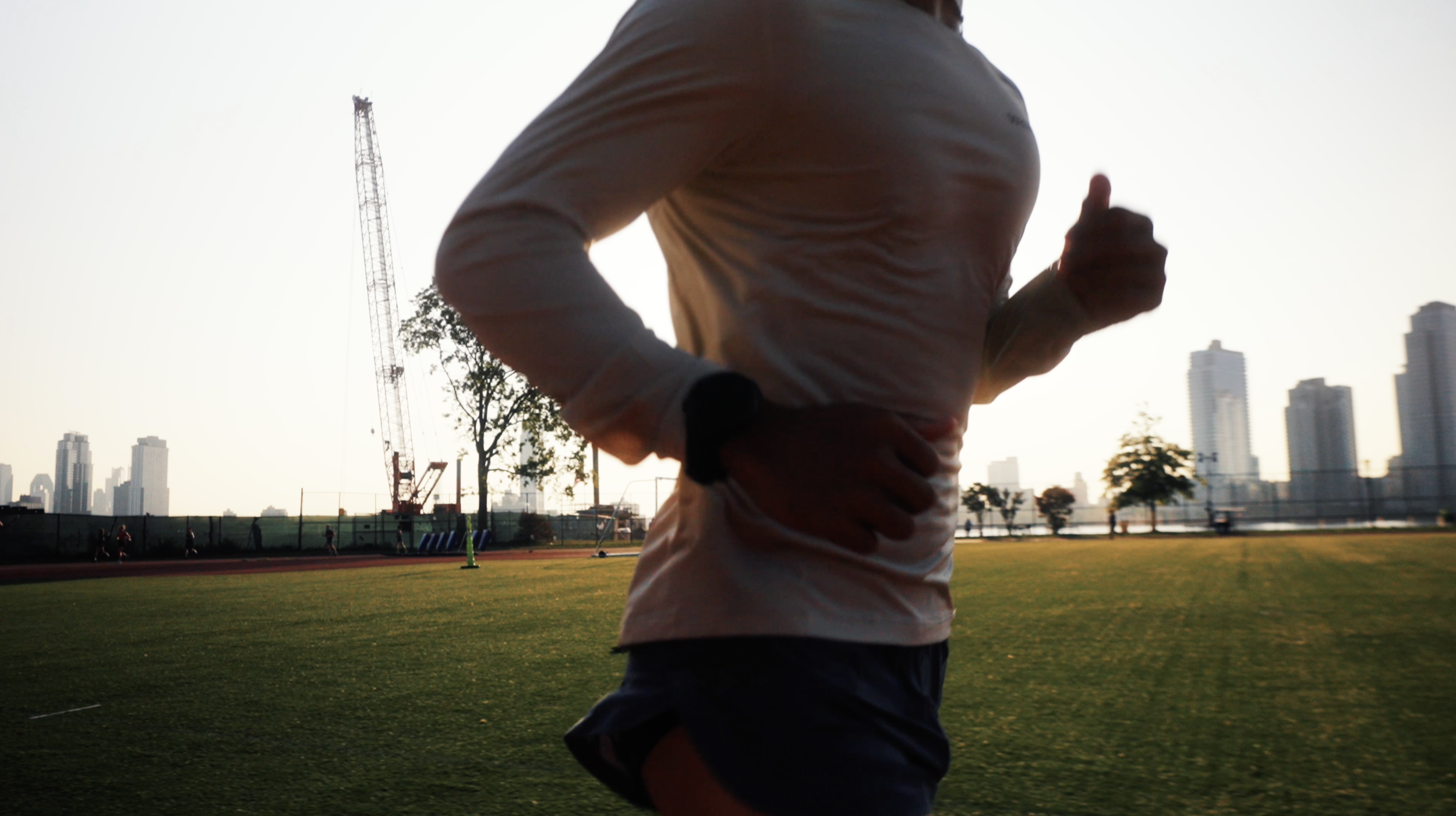 A person jogging on a running track at a park or sports field with a city skyline and tall buildings in the background during sunset or sunrise.