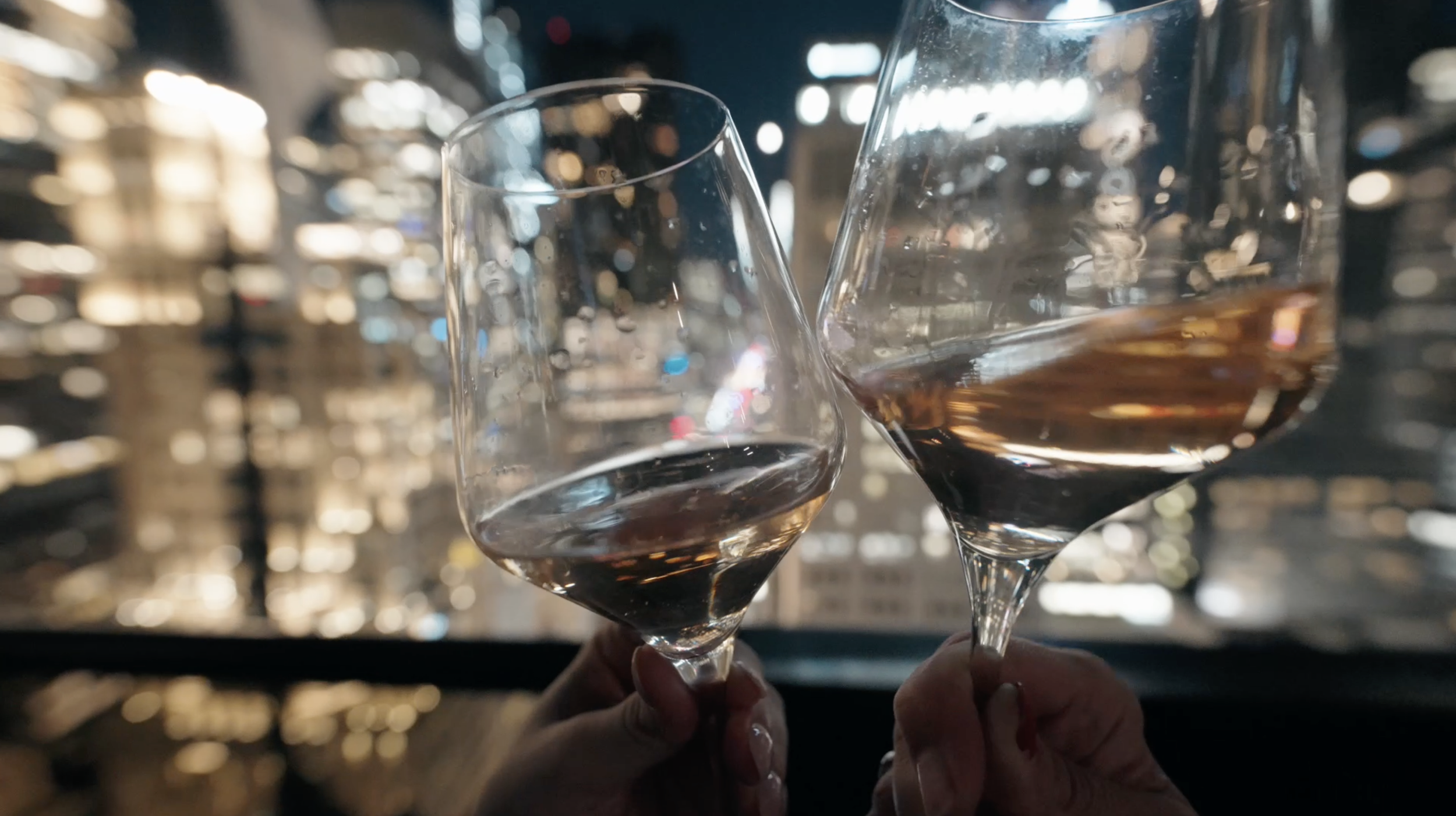 Two glasses of rosé wine being clinked together in a toast, with a blurred cityscape background at night.