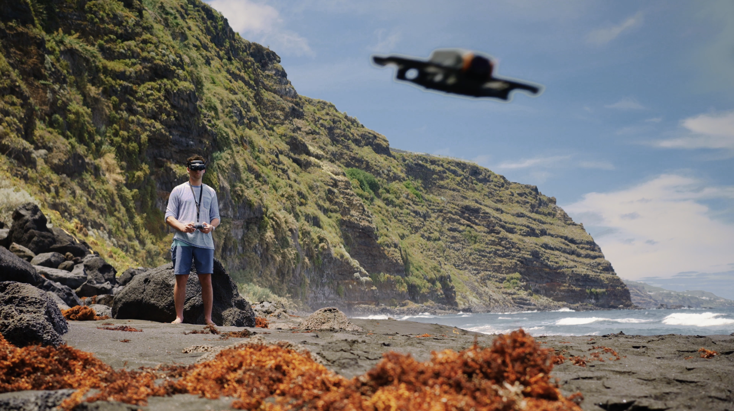 A person standing on a rocky beach at the base of green mountains, flying a drone with a remote control, under a partly cloudy sky.