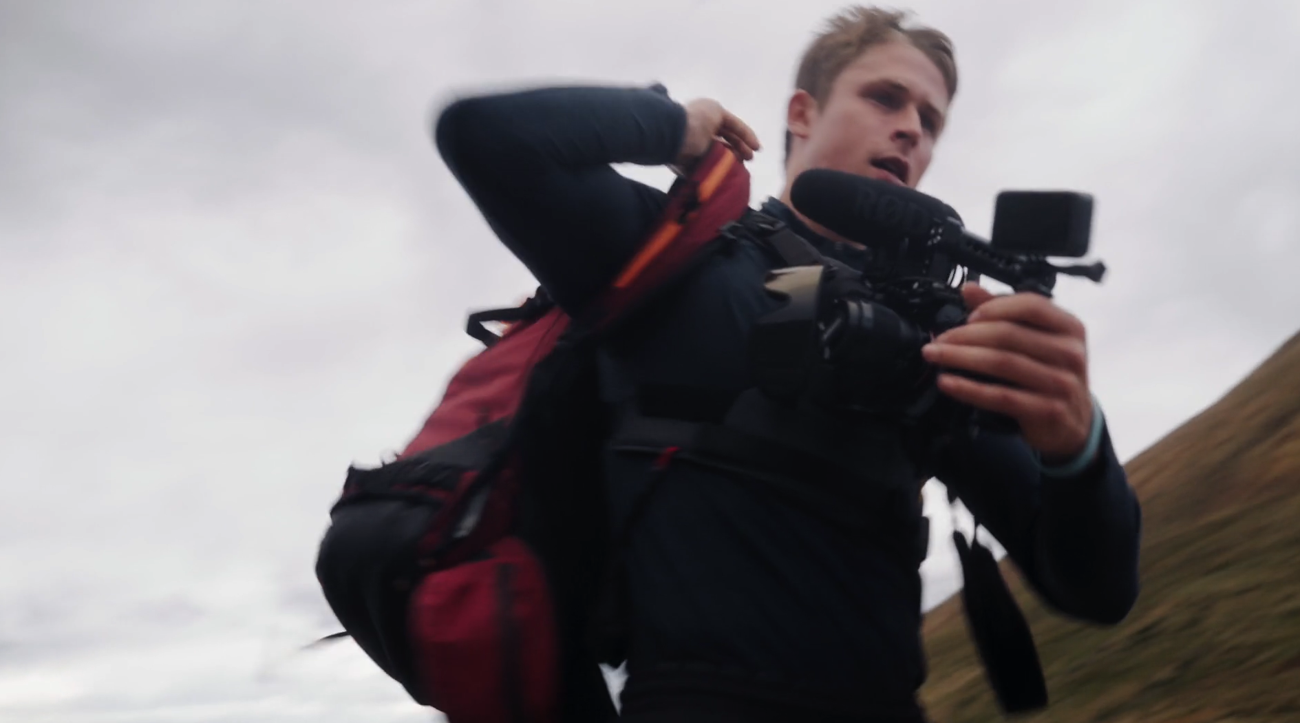 Young man with a backpack filming with a camera outdoors on a cloudy day.
