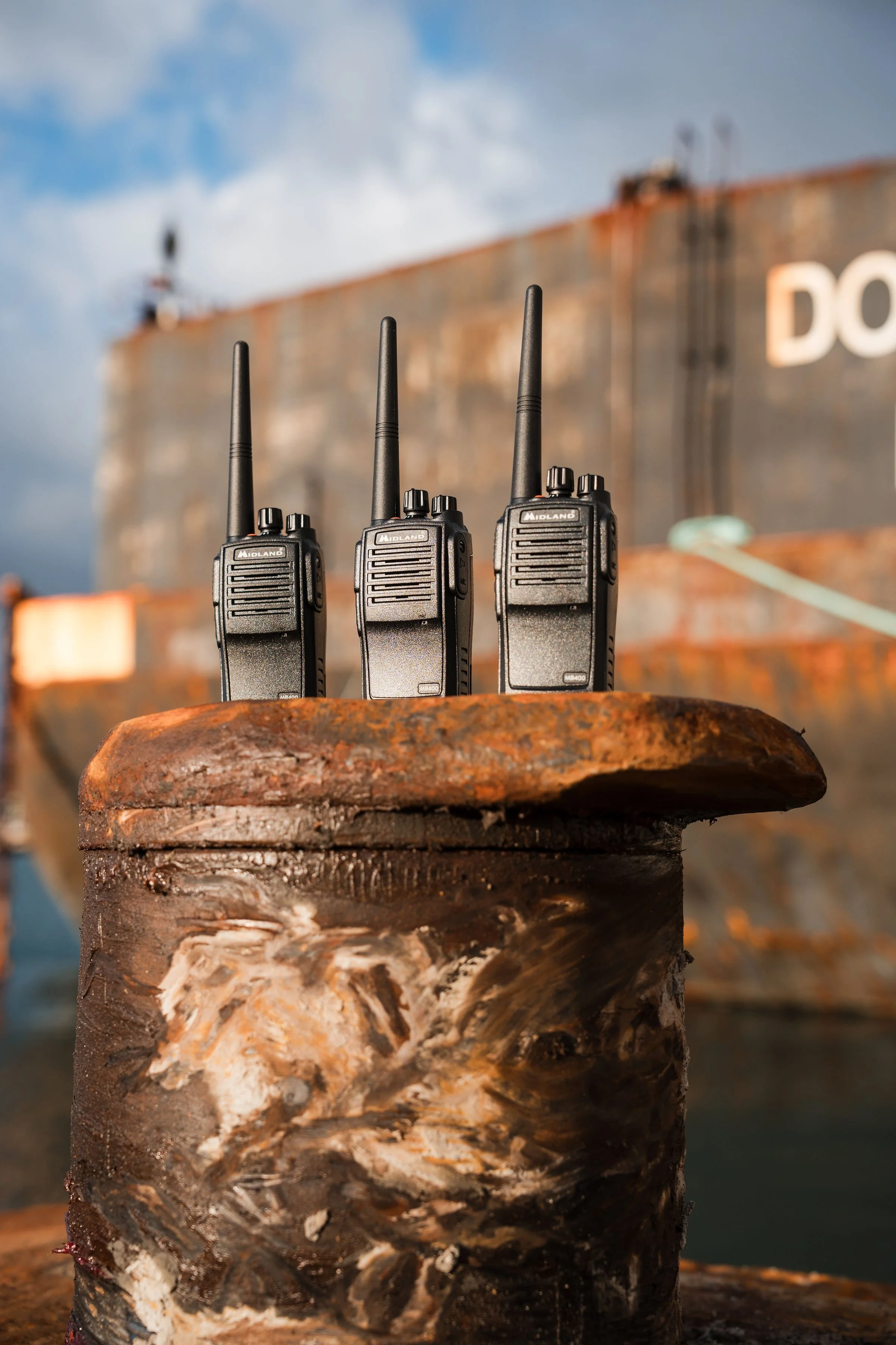 Three walkie-talkies placed on a rusty metal surface with a ship in the background and a partly cloudy sky.