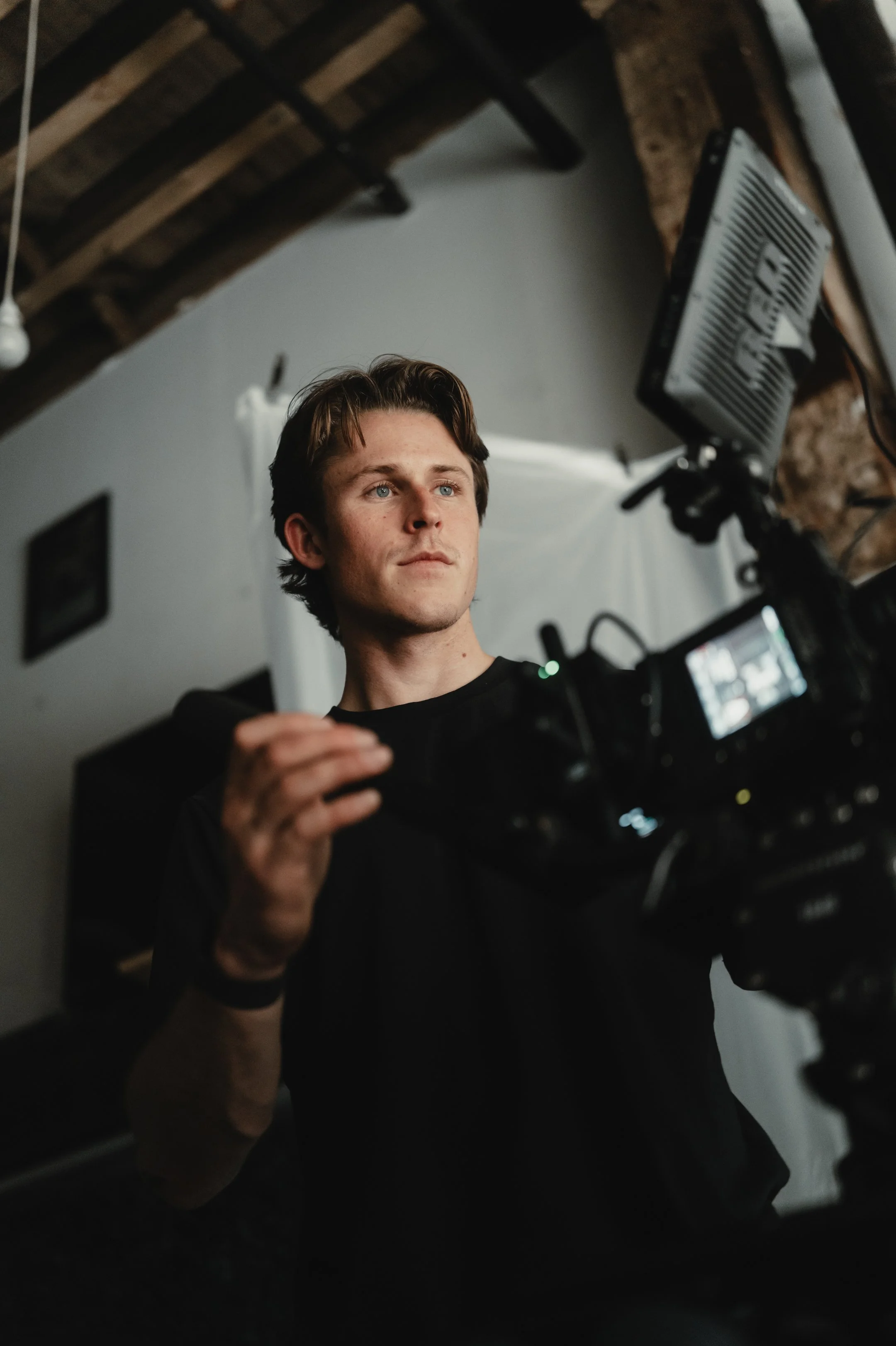 A man operating a professional camera in a photography studio with a white backdrop and exposed ceiling beams.