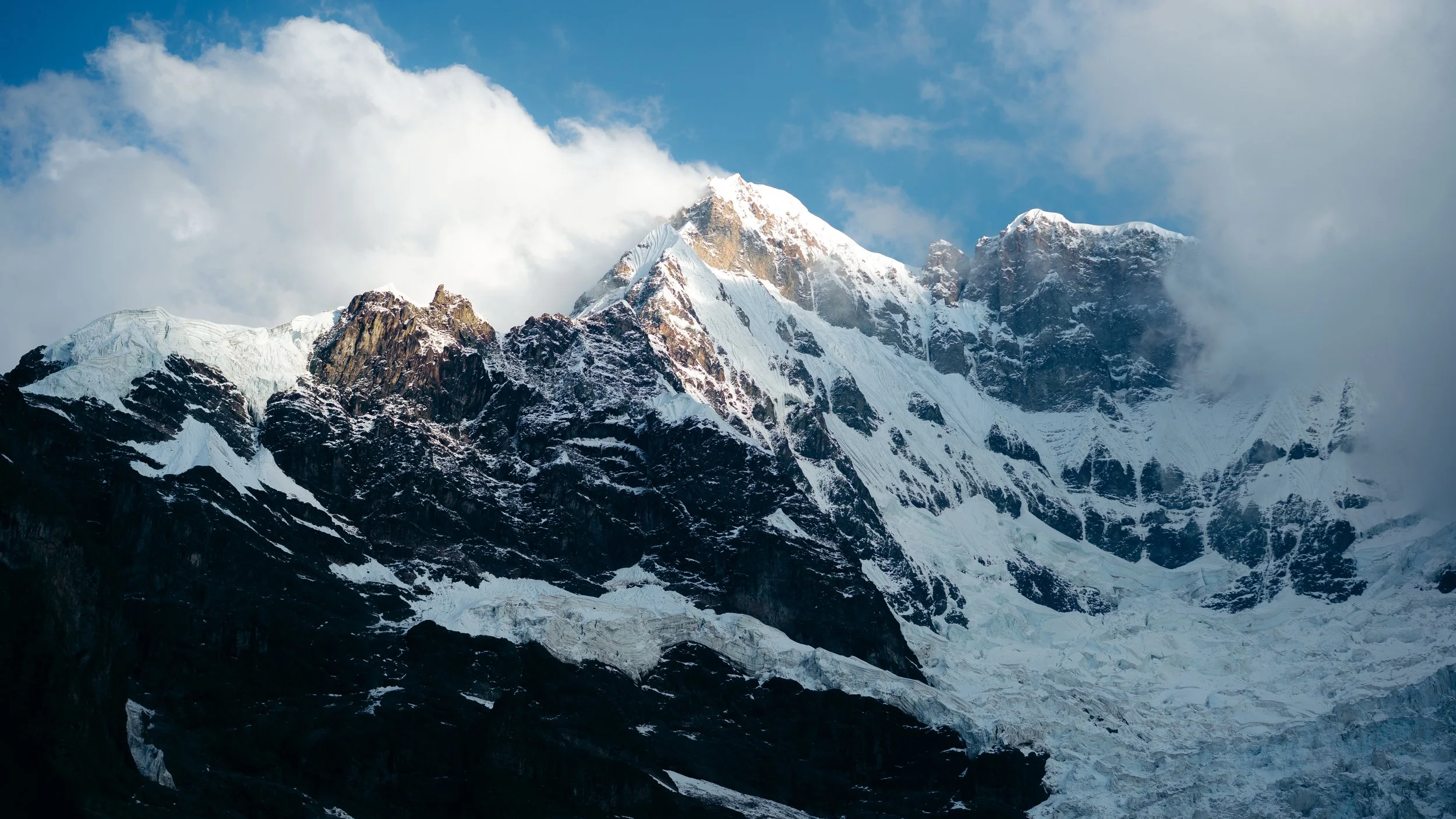 Snow-capped mountain peaks with rocky cliffs and a glacier, partly covered by clouds, under a blue sky.
