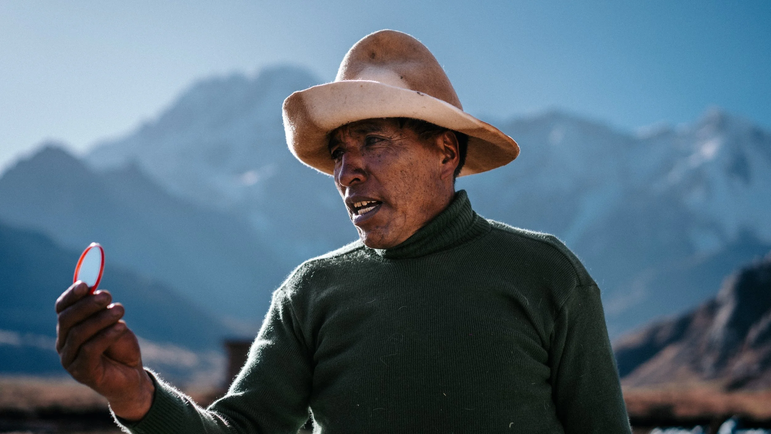 A man wearing a large beige hat and a green sweater holding a small mirror, with mountains in the background.