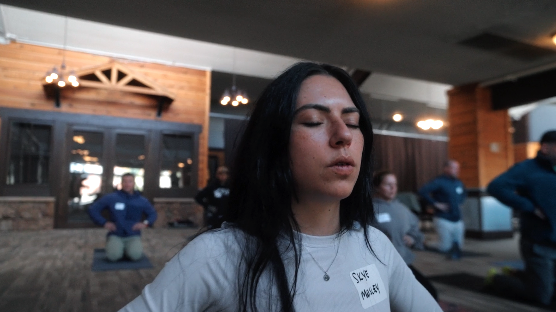 A woman with black hair, closed eyes, wearing a white shirt with a name tag that reads 'Skye Molly', sitting outdoors during a meditation or yoga session at a rustic venue with wooden walls and warm lighting.
