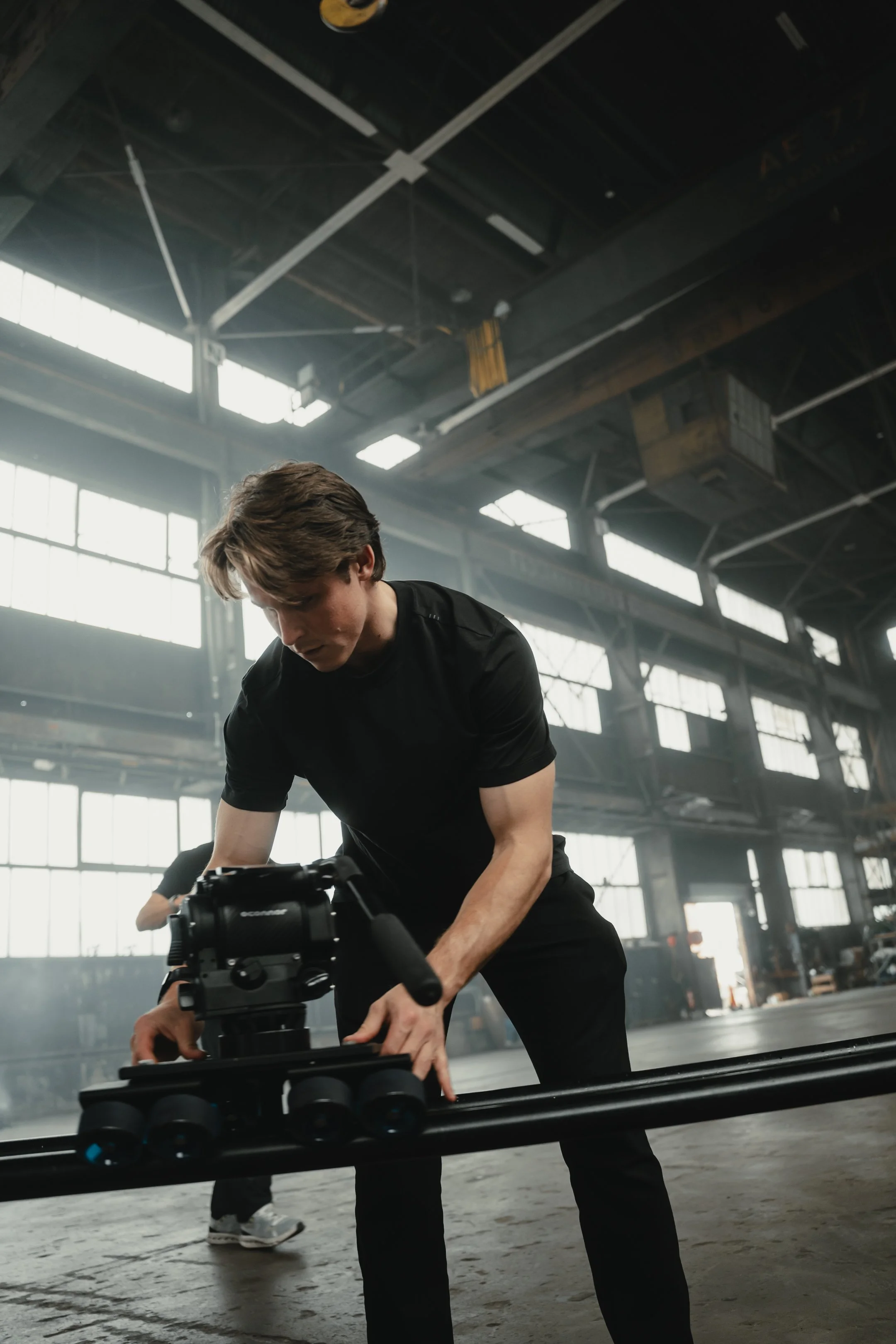 A young man in a black t-shirt and pants adjusting camera equipment on a dolly inside an industrial warehouse with large windows and high ceilings.