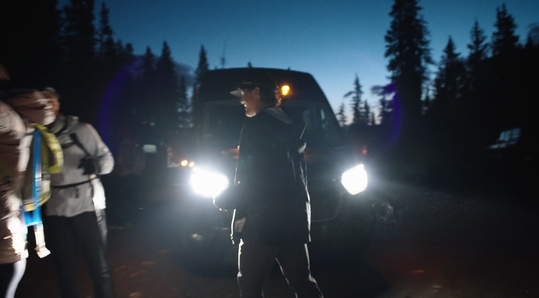 Three people outdoors at dusk near a black van with headlights on, surrounded by tall trees.