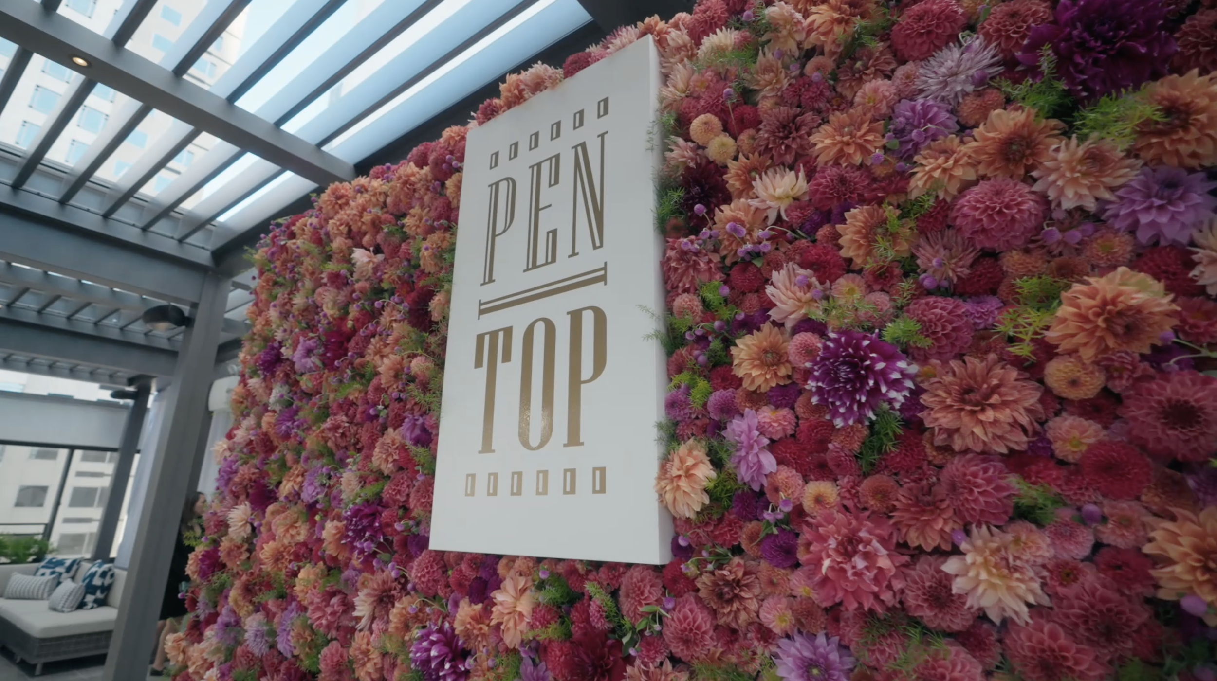 A floral wall with a white sign that reads "PEN TOP" in gold letters, located on a balcony or rooftop with a glass roof and city buildings in the background.