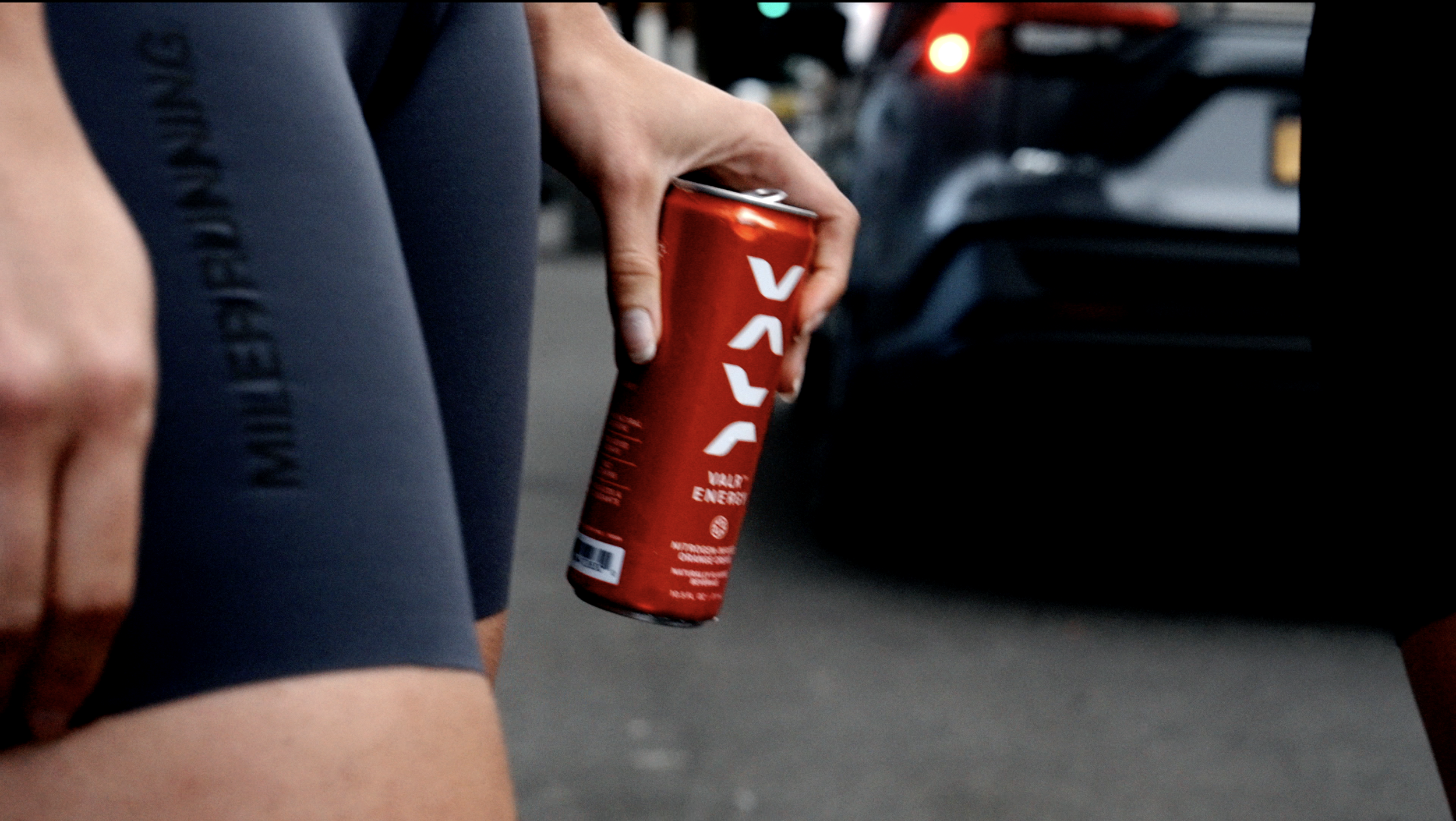 A person holding a red energy drink can at a race or outdoor event, wearing tight black athletic shorts.
