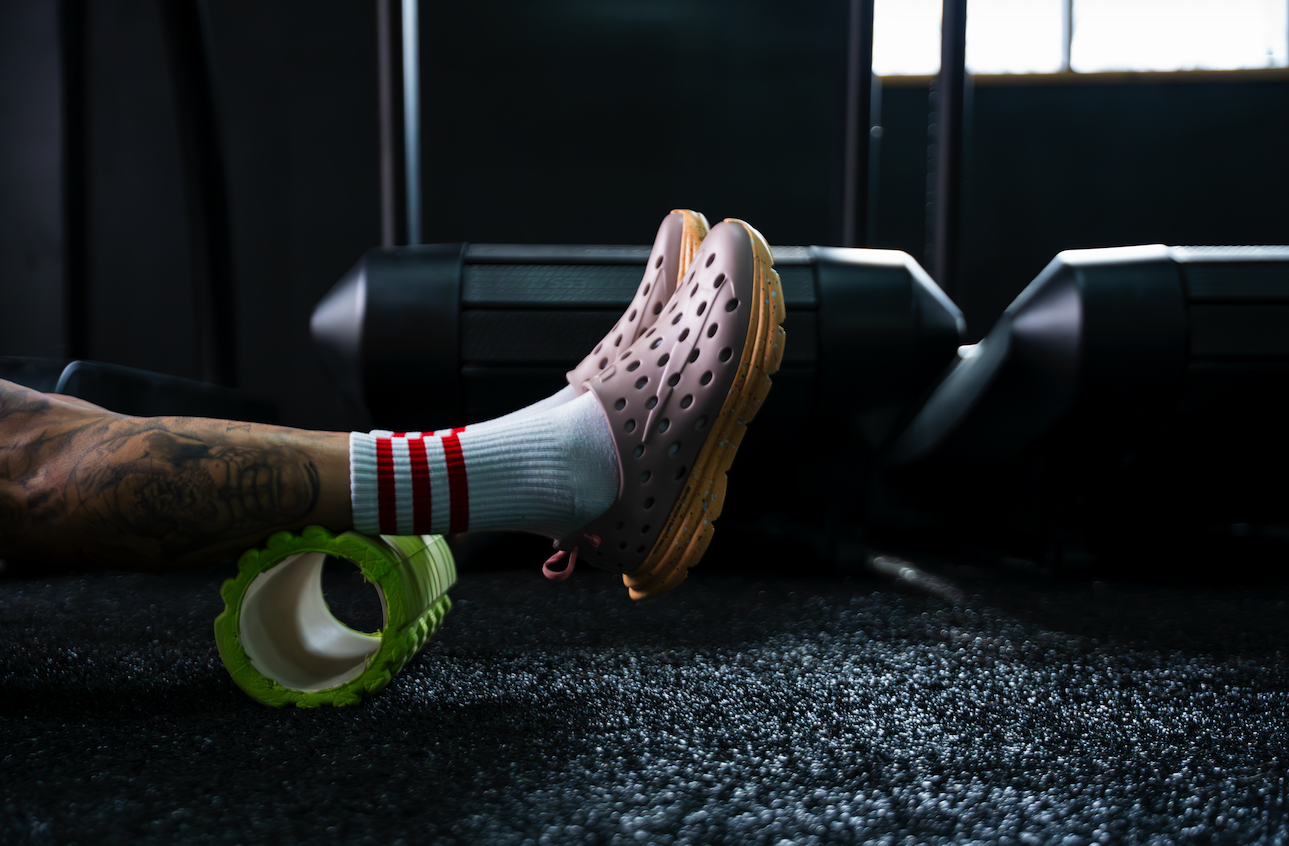 Person's legs with tattoos, wearing white socks with red stripes and gray Crocs, resting on a black exercise mat near treadmill at gym.