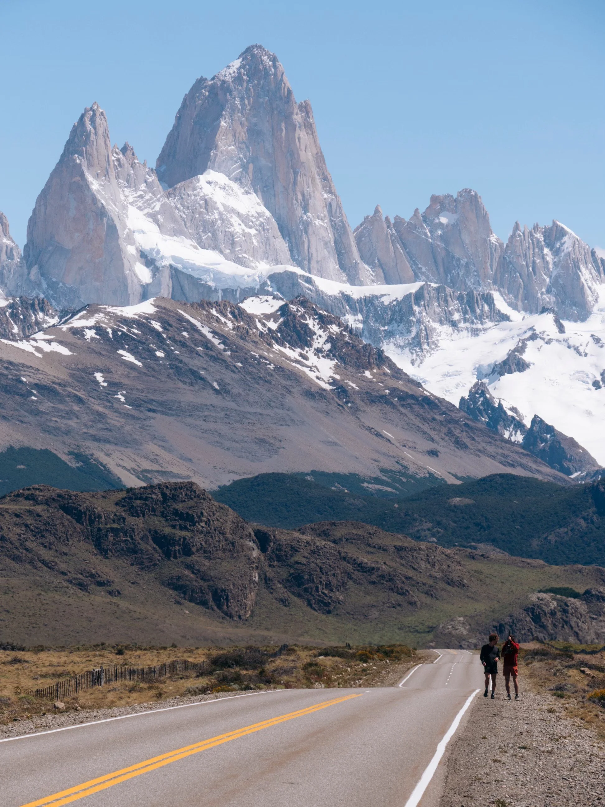 Two people hiking on a road near mountains with snow-capped peaks in the background.