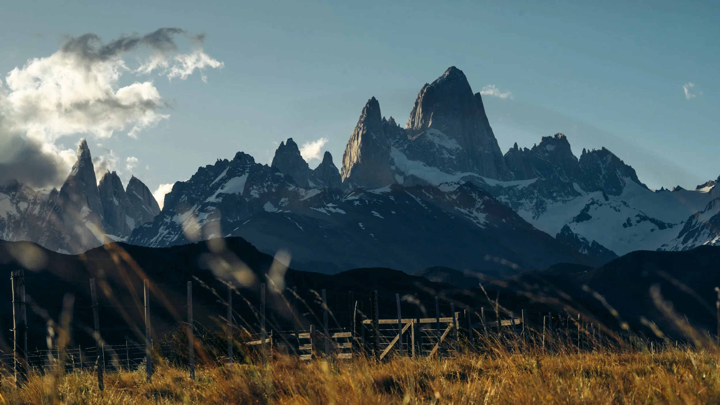 Snow-capped mountains under a partly cloudy sky with a fence and grassy foreground.