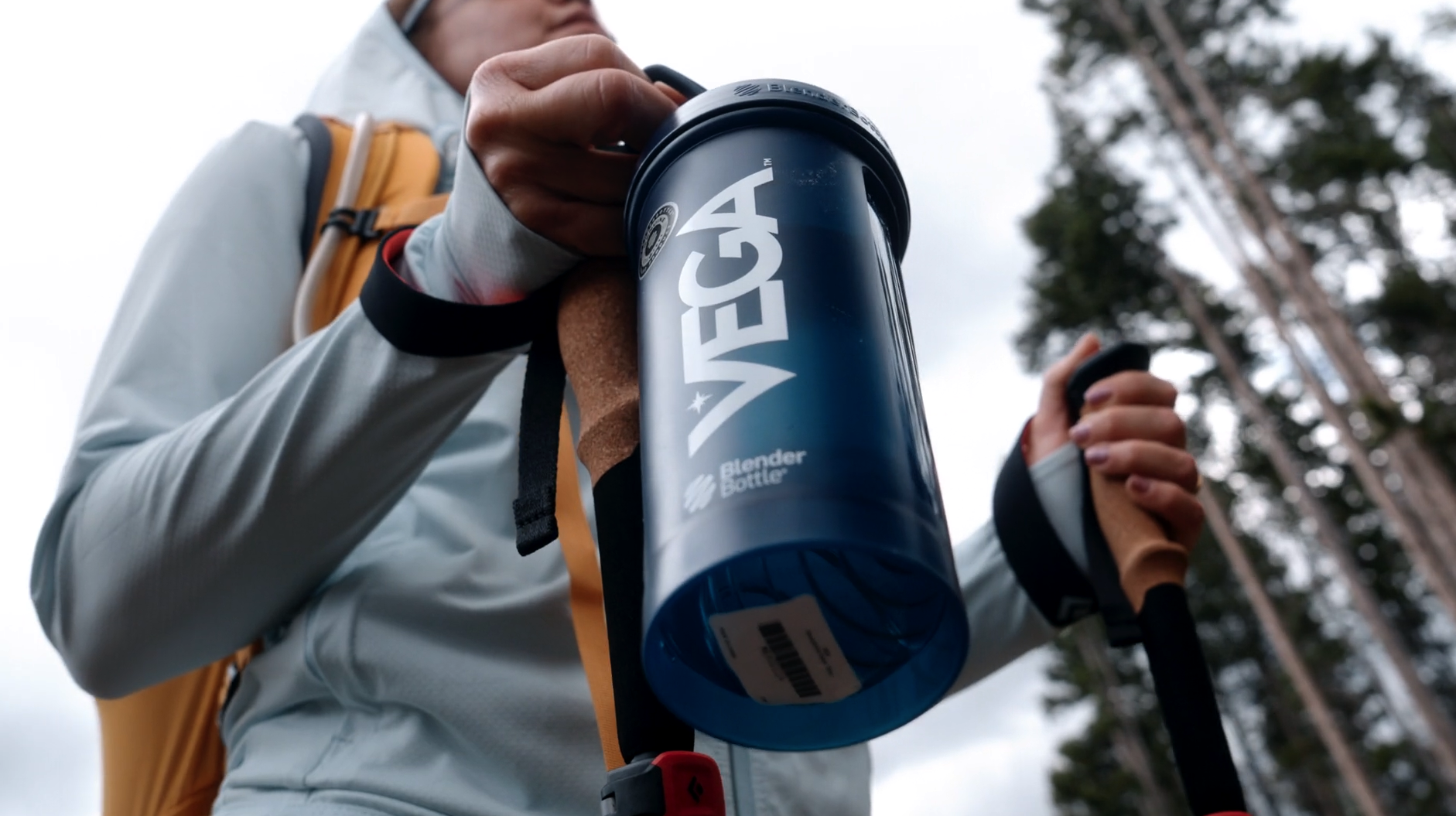 Close-up of a person holding an insulated blue water bottle with the brand name 'VEGA' on it, outdoors with trees visible in the background.