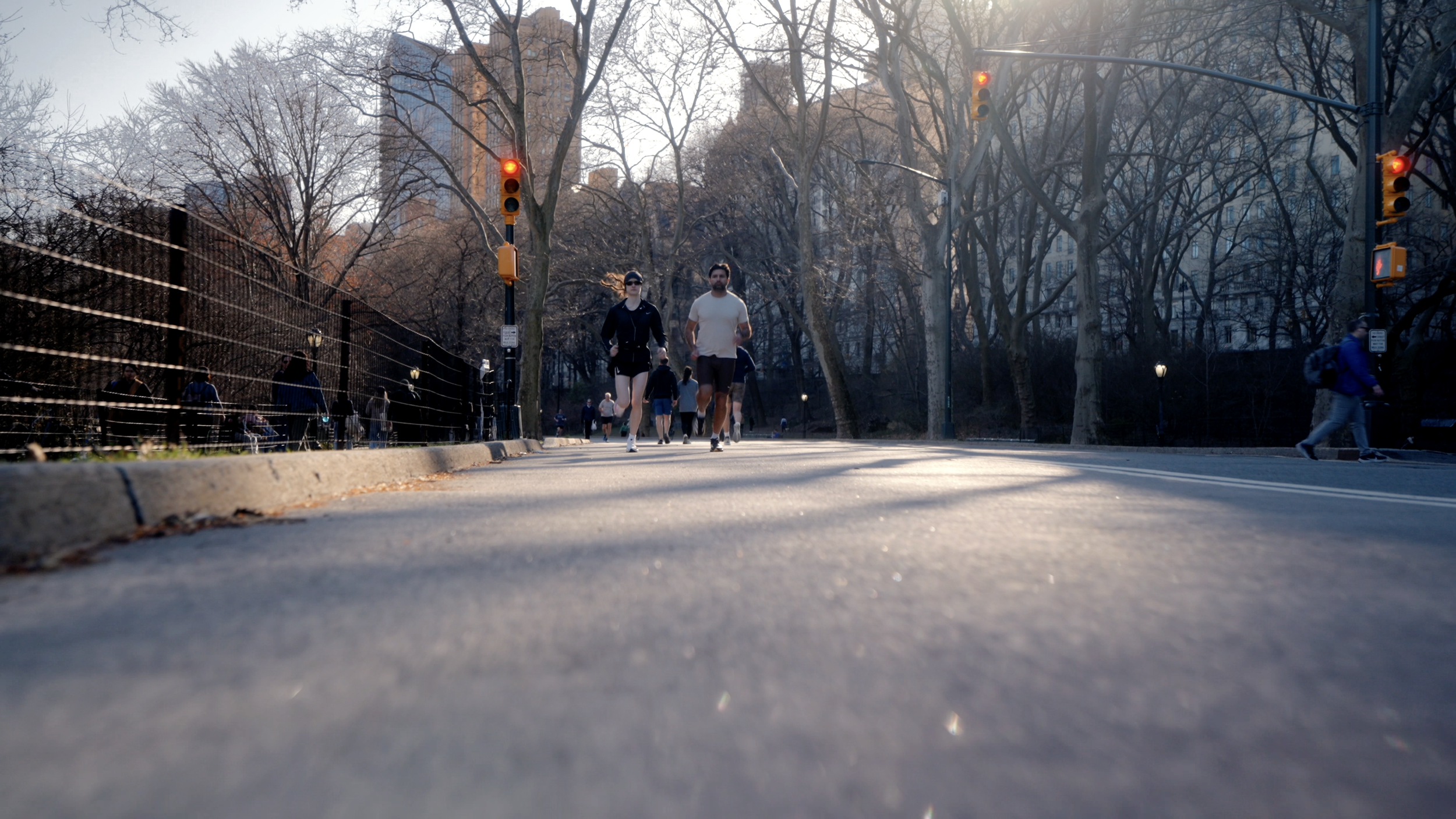 People jogging and walking in a park on a city street with traffic lights and leafless trees during a sunny day.