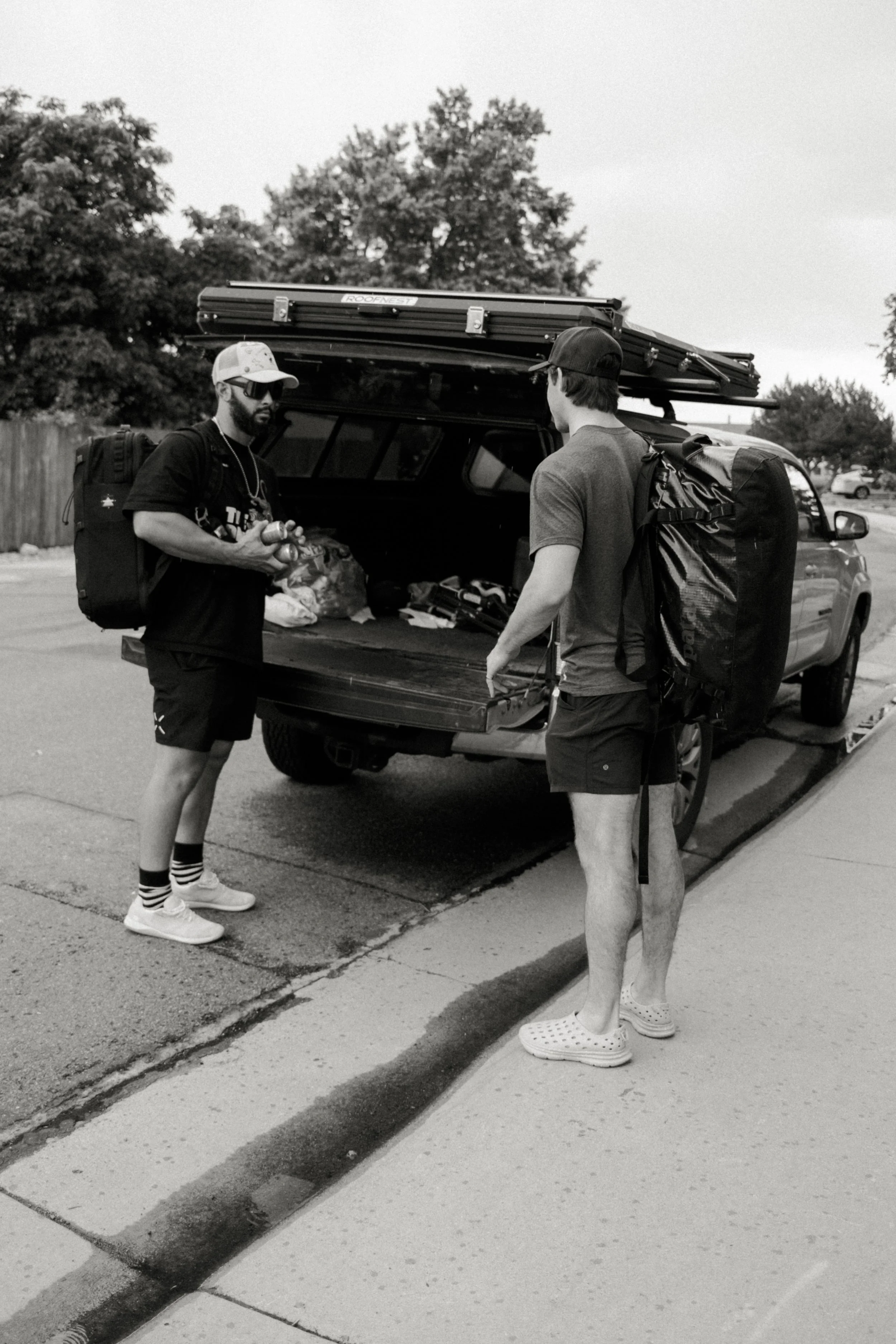 Two men with backpacks and hats loading gear into the back of a pickup truck parked on the side of a street with trees in the background.