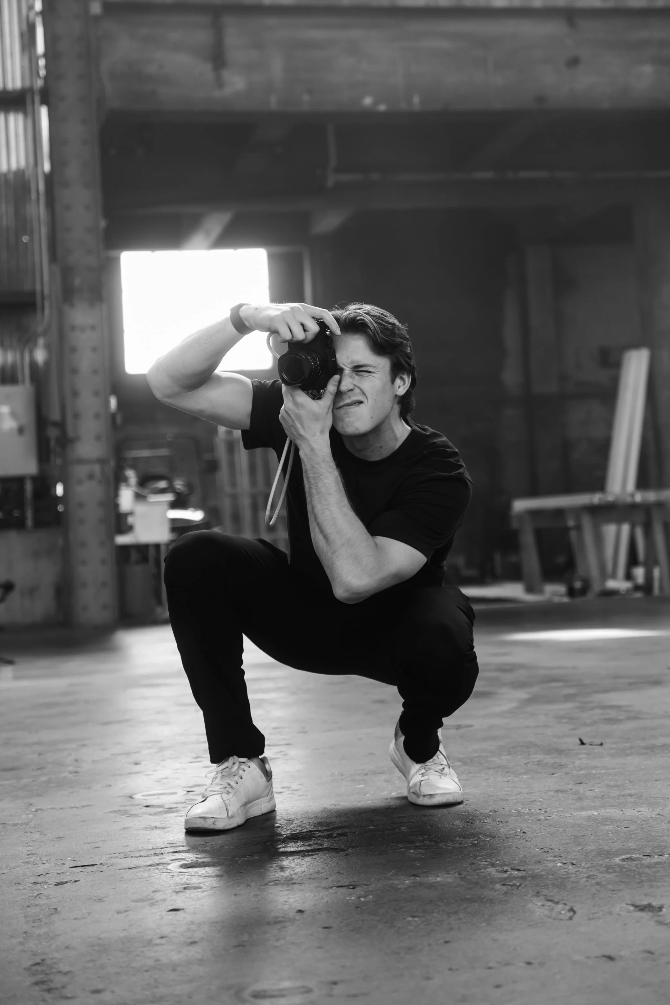 A young man is squatting indoors, holding a camera up to his eye, squinting as he looks through the viewfinder. He is wearing a black t-shirt, black pants, and white sneakers, with a background of a warehouse or workshop with wooden beams and equipme