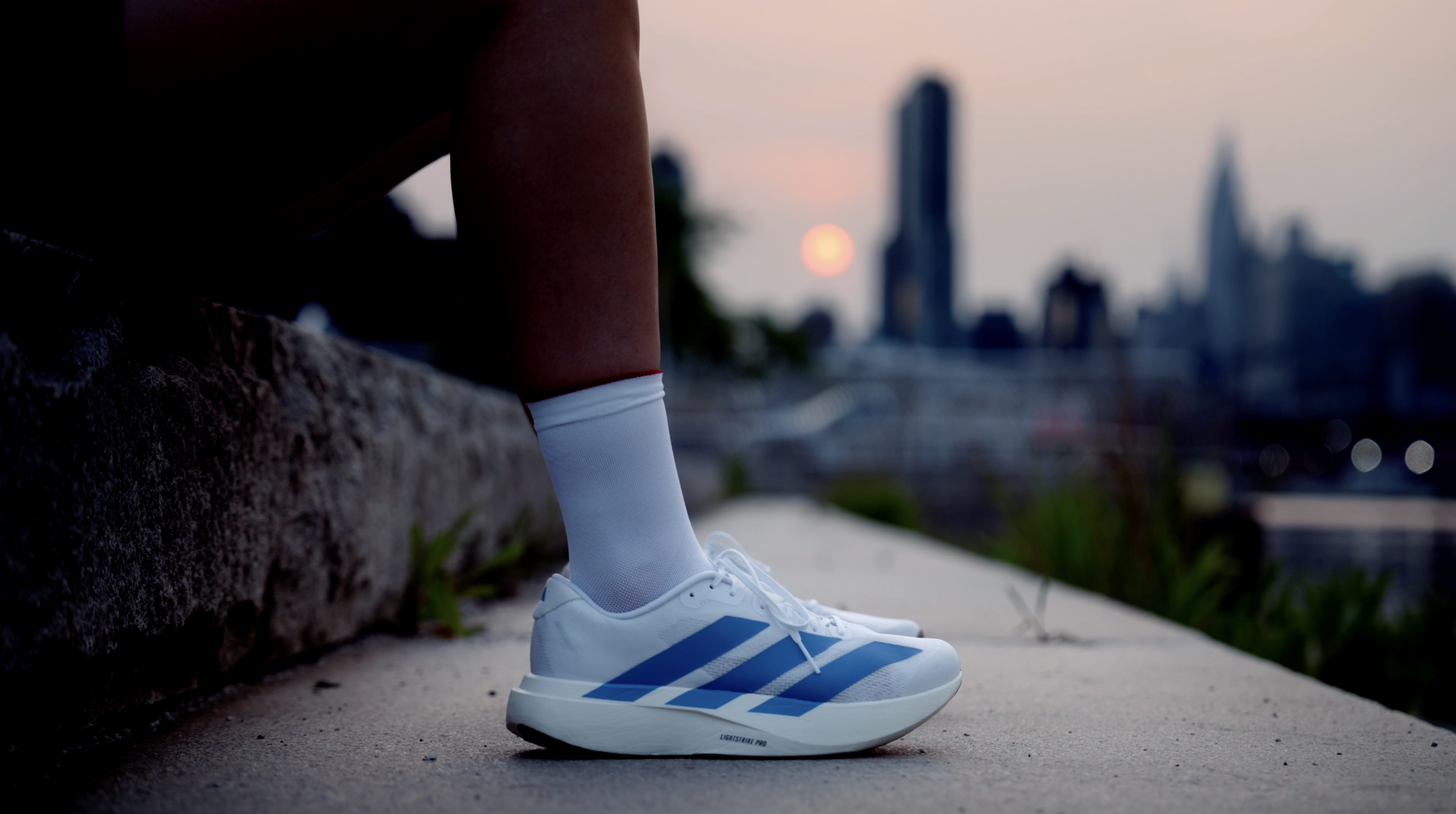 Close-up of a person wearing white athletic shoes with blue stripes and white socks, standing on a concrete surface with a city skyline and sunset blurred in the background.