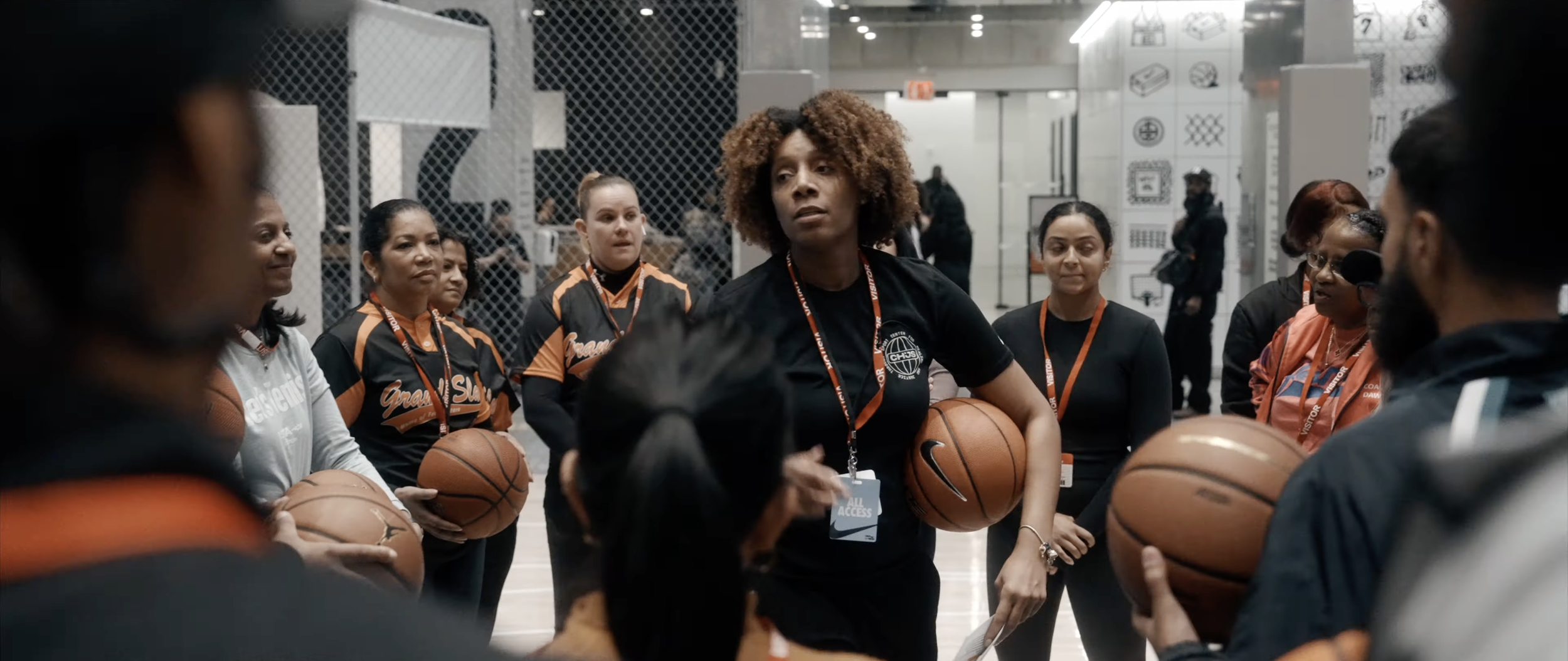 A group of women wearing sports jerseys and holding basketballs, gathered around a woman with curly hair giving instructions in an indoor sports facility.