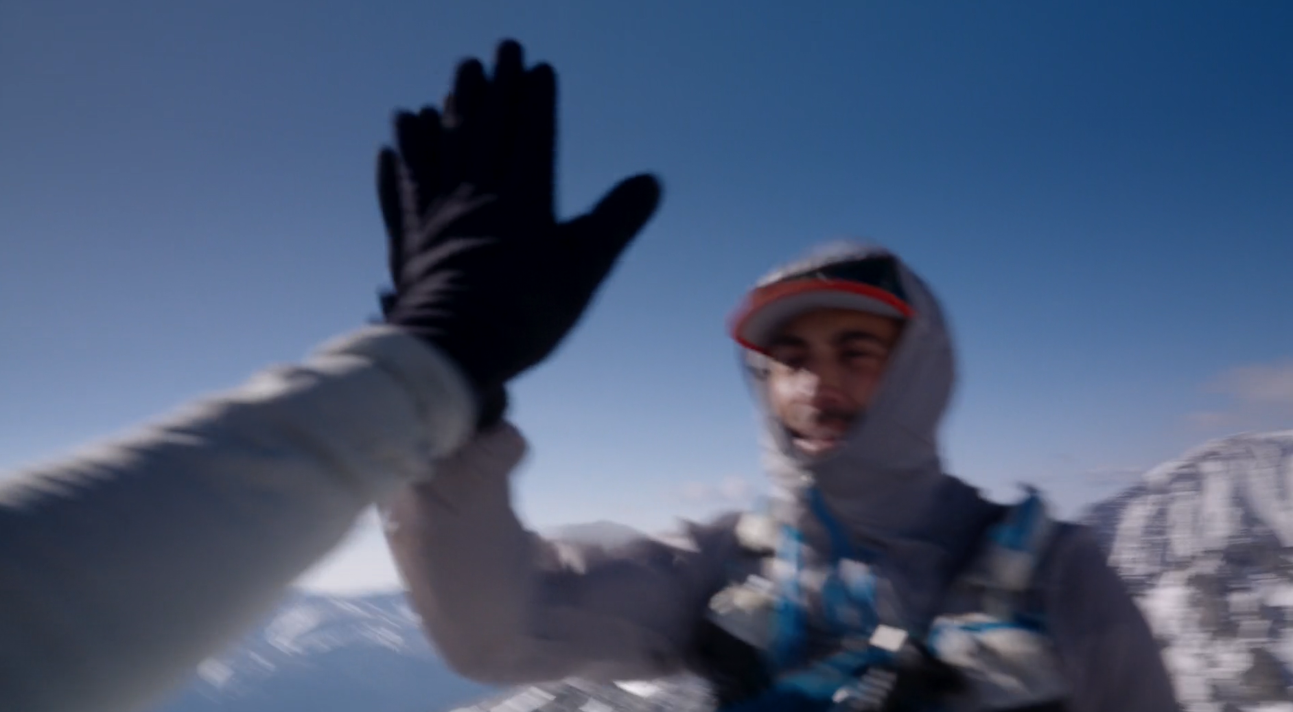 A person in winter gear high-fives another person outdoors against a clear blue sky with snowy mountains in the background.