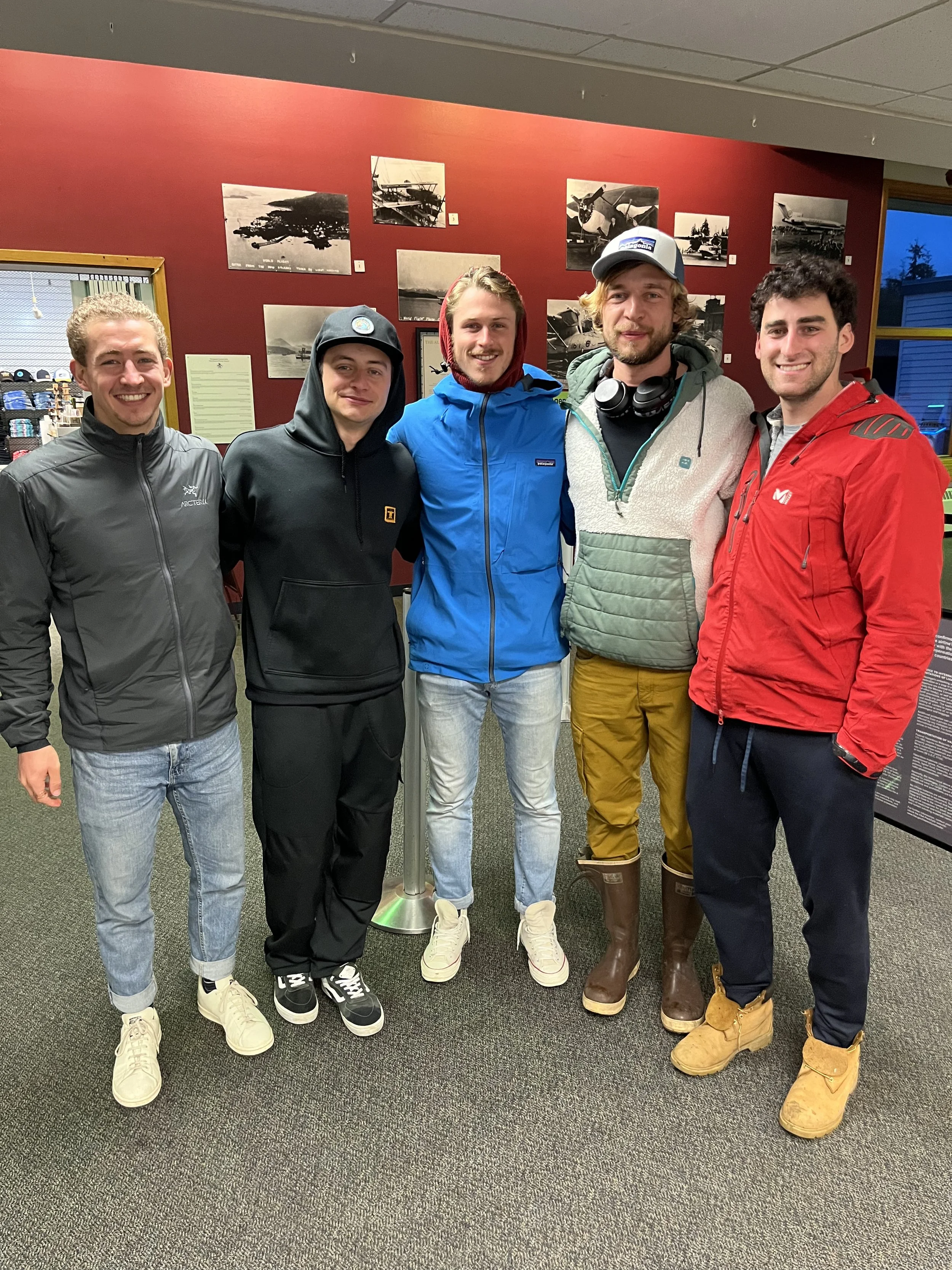 Group of five young men standing inside, dressed in casual and outdoor winter clothing, in front of a red wall with black and white photos.