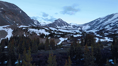 Snow-covered mountains and pine trees in a valley under a cloudy sky. An XPT Commercial