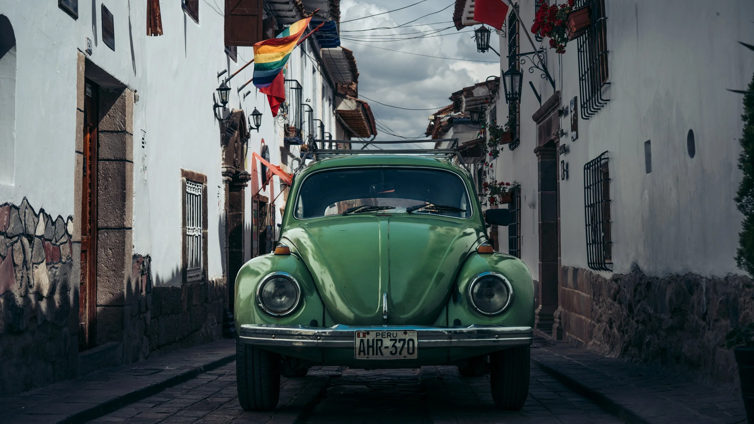 A vintage green Volkswagen Beetle parked on a cobblestone street in a historic neighborhood with white colonial-style buildings and balconies decorated with flowers.