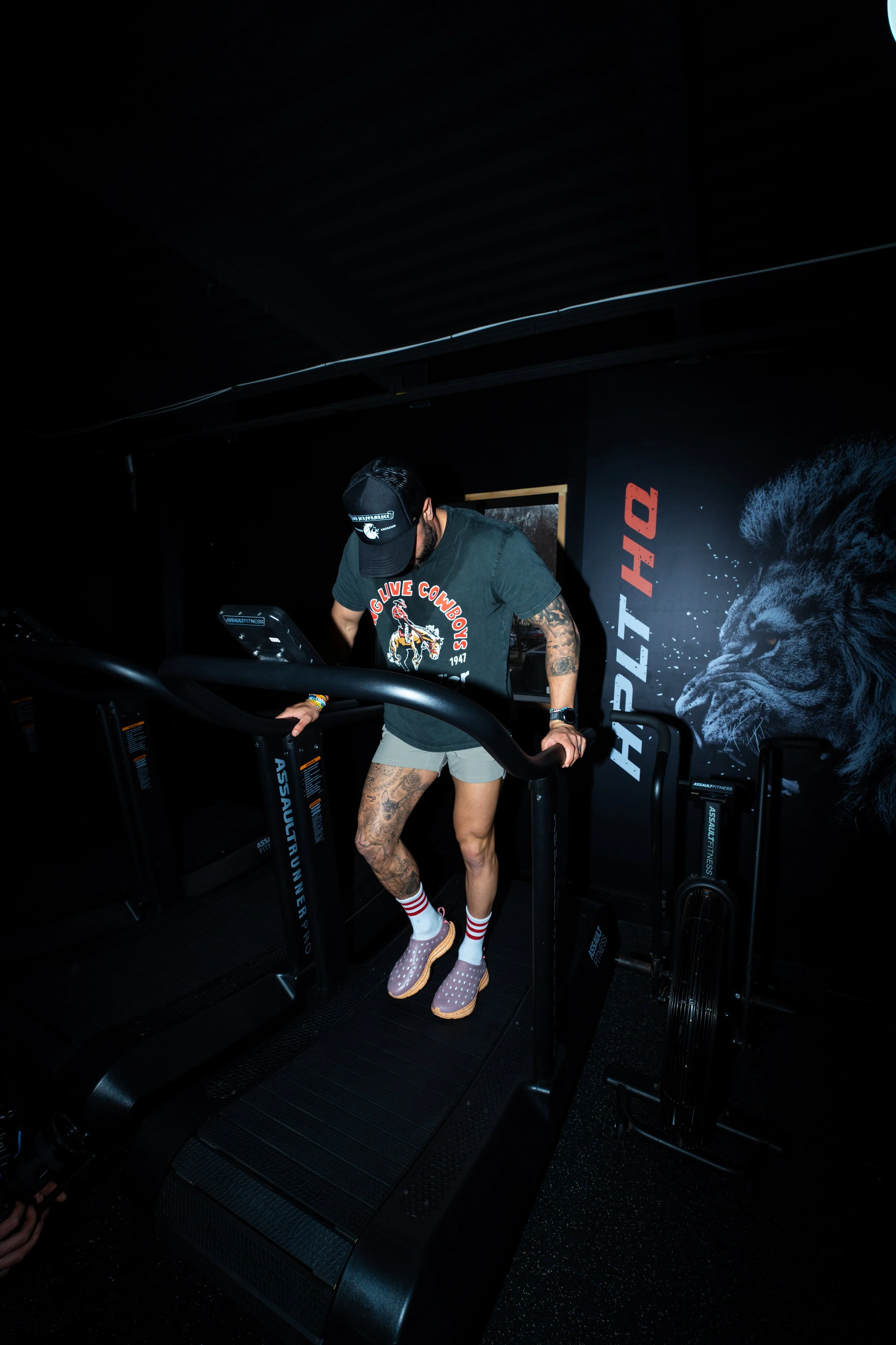 A man in a black cap, black t-shirt, gray shorts, and pink Crocs is walking on a treadmill in a dark gym with a black lion mural.