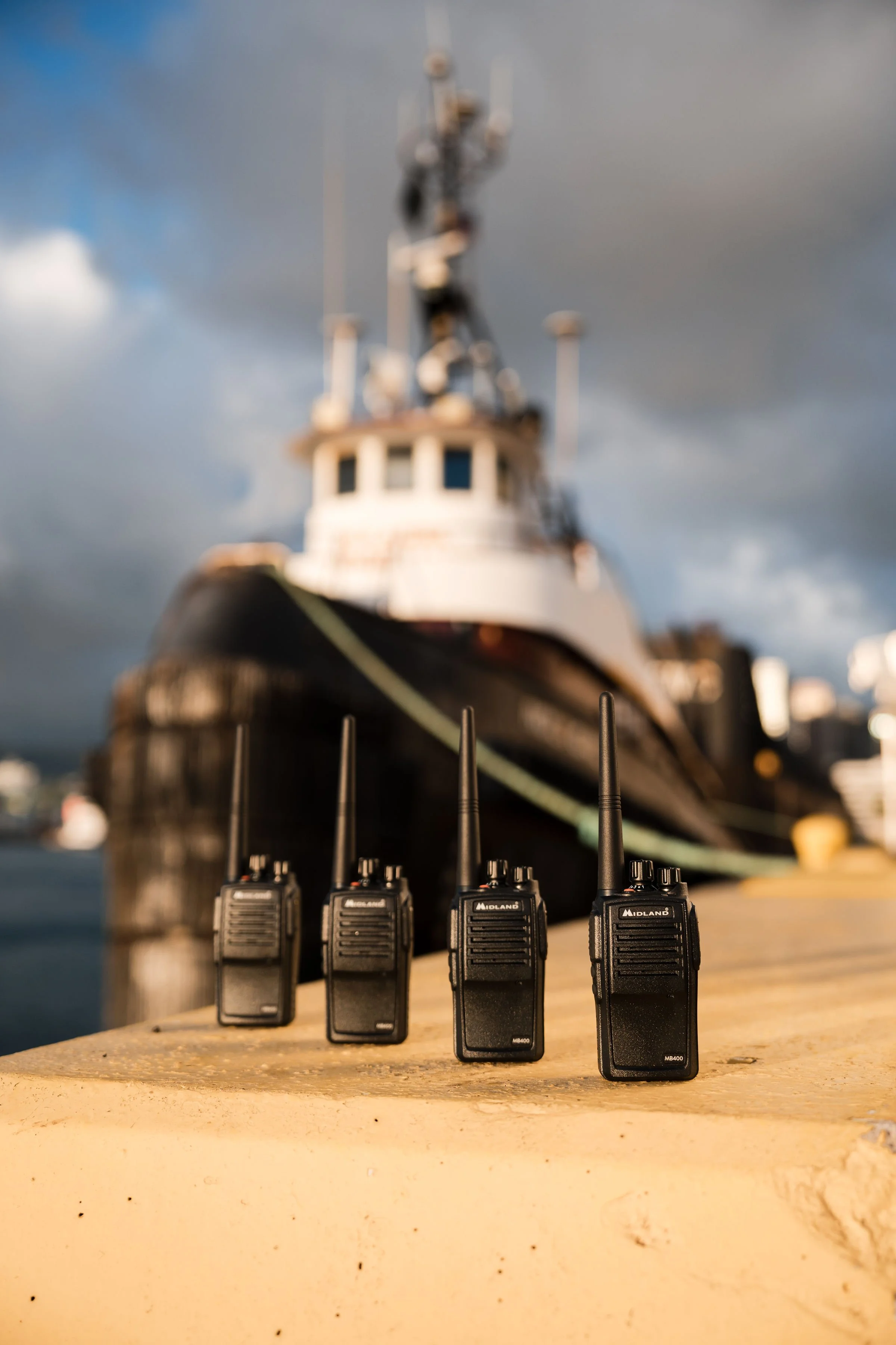 Four walkie-talkies lined up on a wooden surface with a large ship docked in the background on a cloudy day.