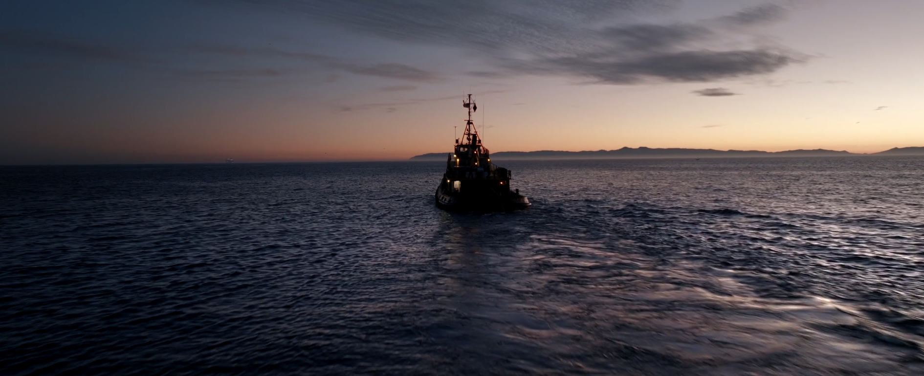 A boat sailing on the ocean during sunset with a distant landmass on the horizon and the sky illuminated with soft colors.