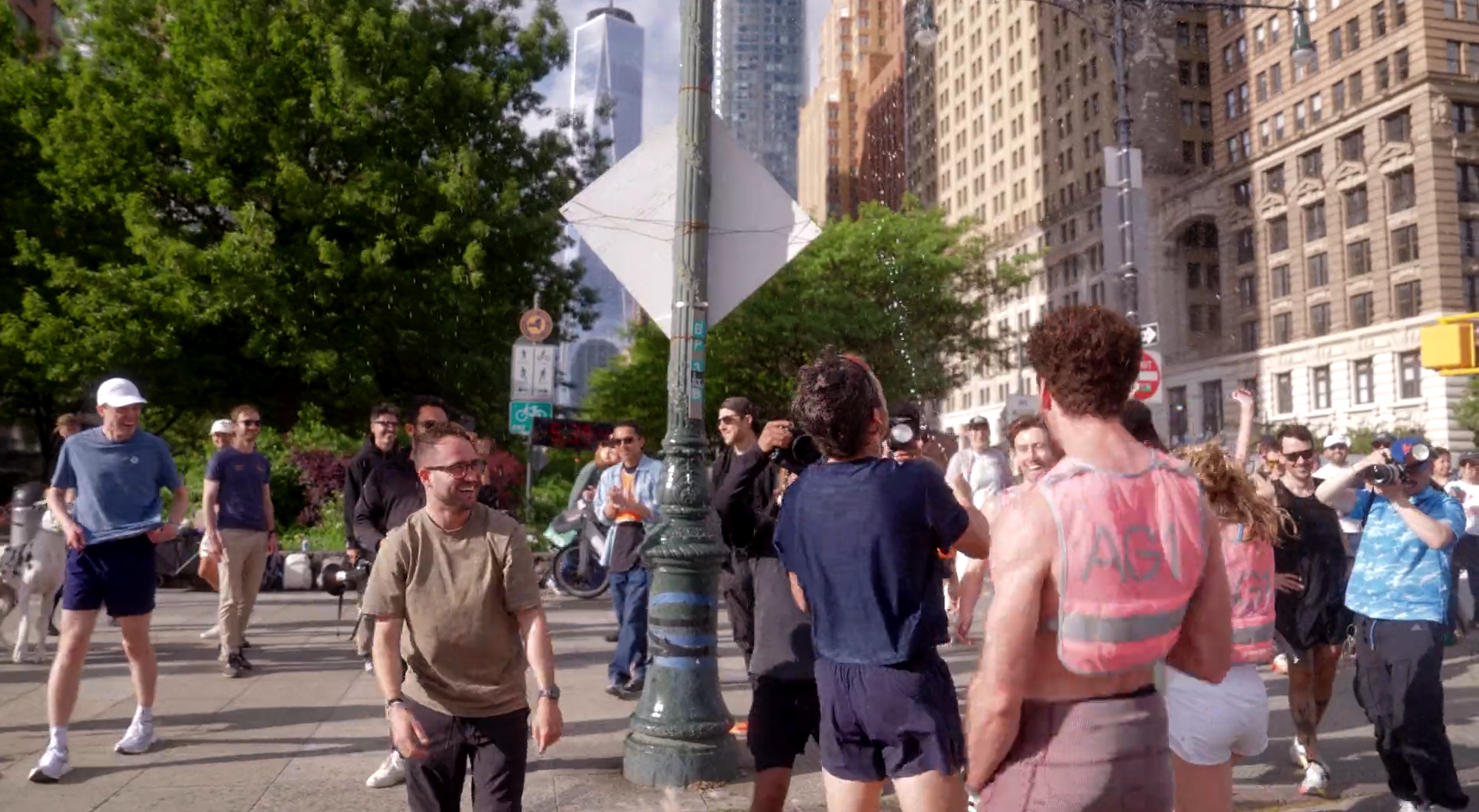 A group of people dancing and laughing outdoors in a city park, with skyscrapers and trees in the background on a sunny day.