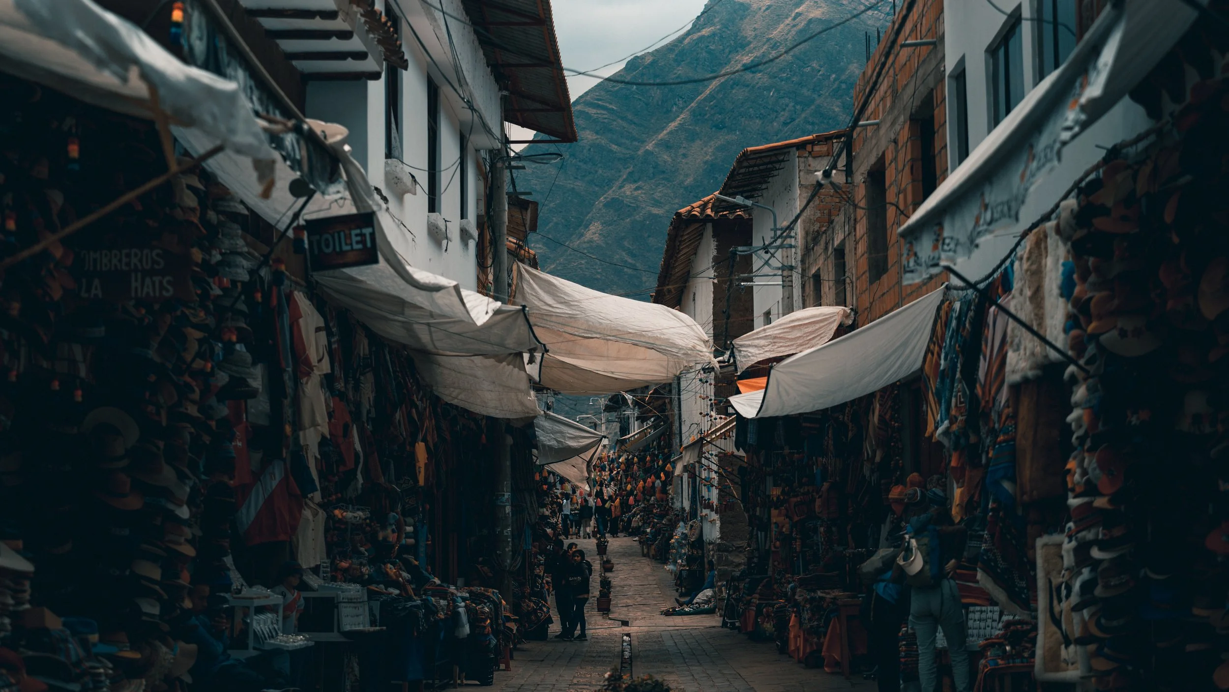 A busy outdoor market street with stalls on both sides, covered in white canopies, with people browsing and mountain scenery in the background.