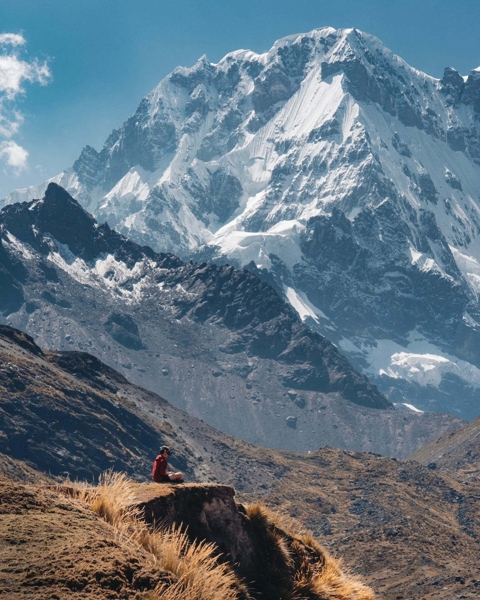 A person sitting on a rock with their legs crossed, wearing a red jacket and headphones, overlooking a mountainous landscape with snow-capped peaks.
