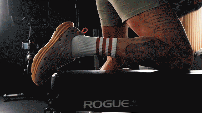 Person with tattoos stretching on a workout bench in a gym, wearing Kane footwear, white socks with red stripes.